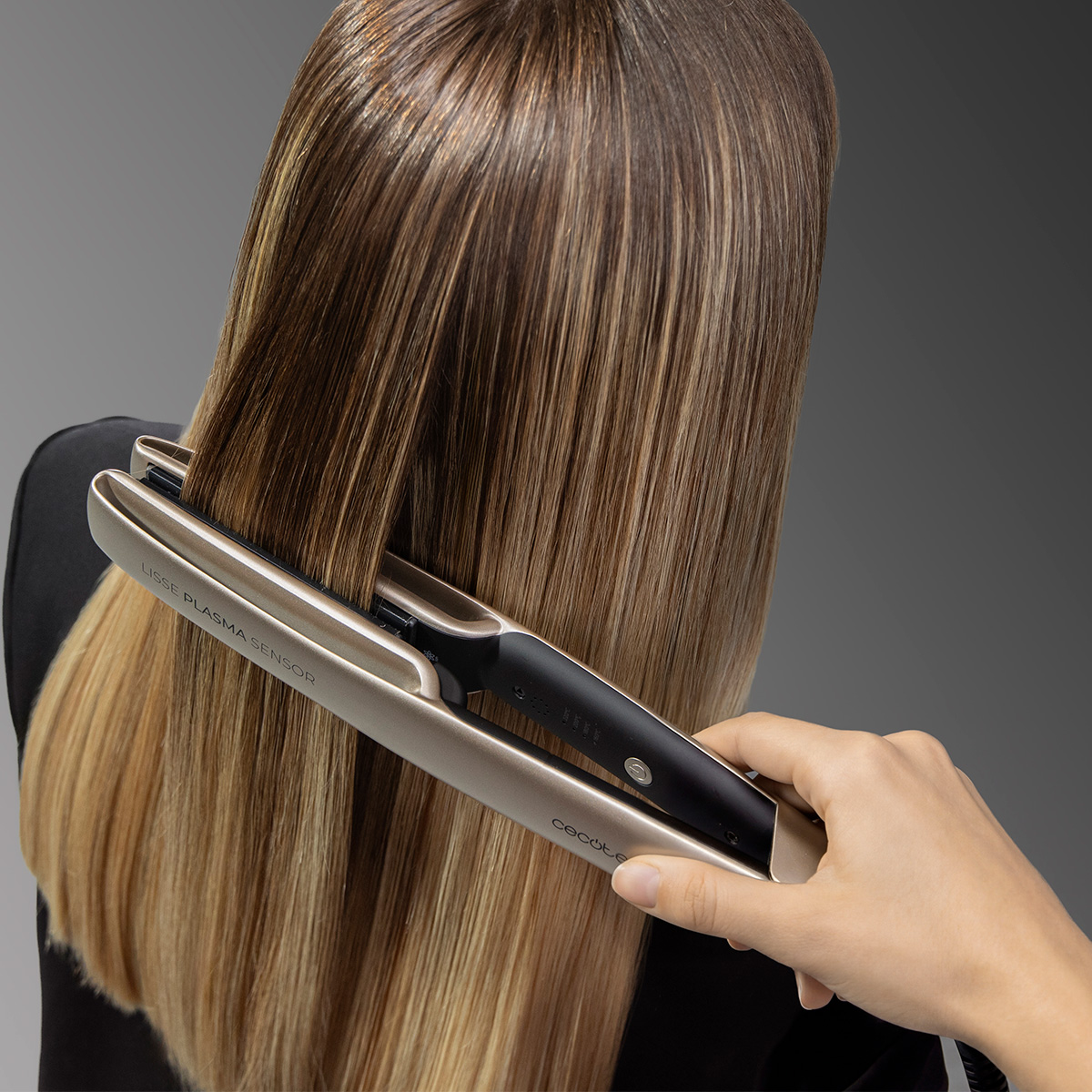 Una persona usa una moderna piastra per capelli oro e nera su capelli castani lunghi e lisci su sfondo grigio.