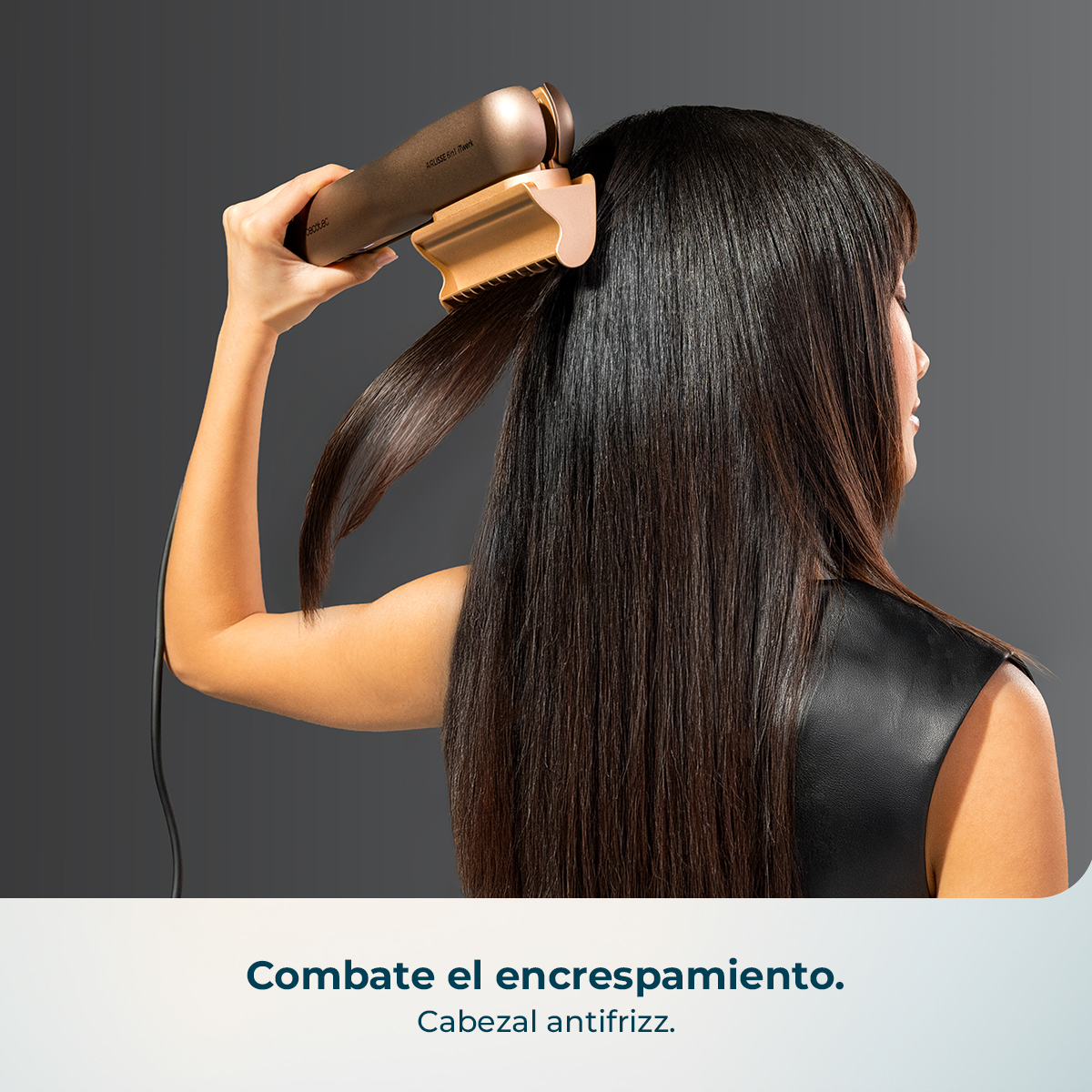 Una mujer moldea su largo cabello oscuro usando una herramienta antifrizz sobre fondo gris.