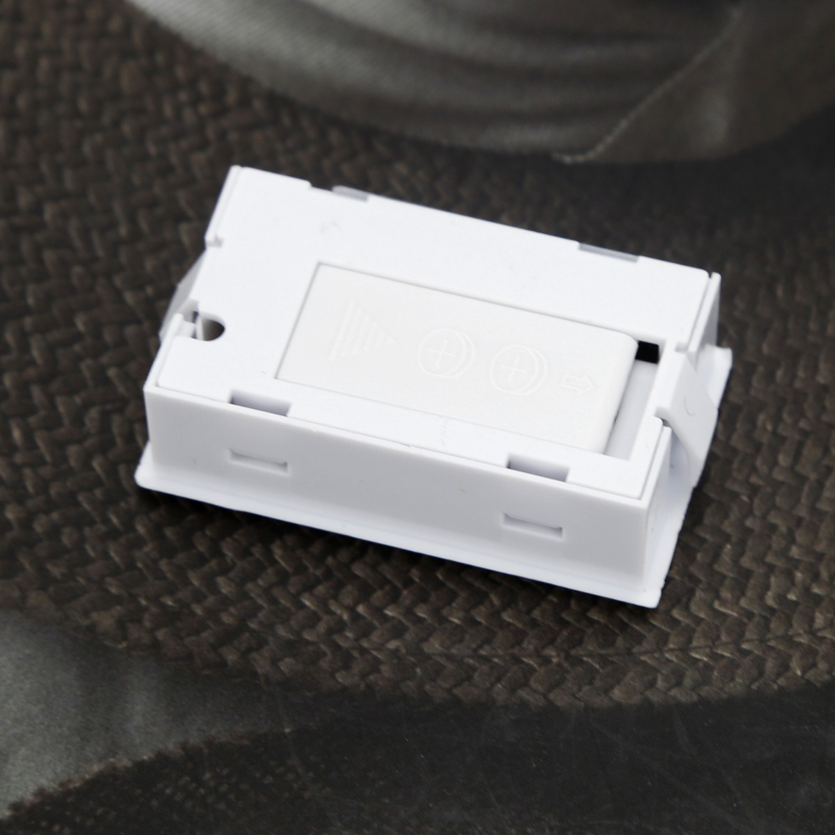 Ein weißes, rechteckiges Kunststoffgehäuse mit einem Batteriefachdeckel auf einer strukturierten Oberfläche.