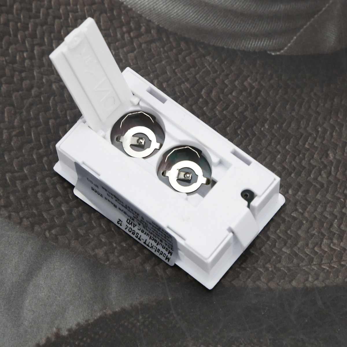 Ein weißes elektronisches Bauteil mit geöffnetem Batteriefach und silbernen Kontakten auf dunklem Untergrund.