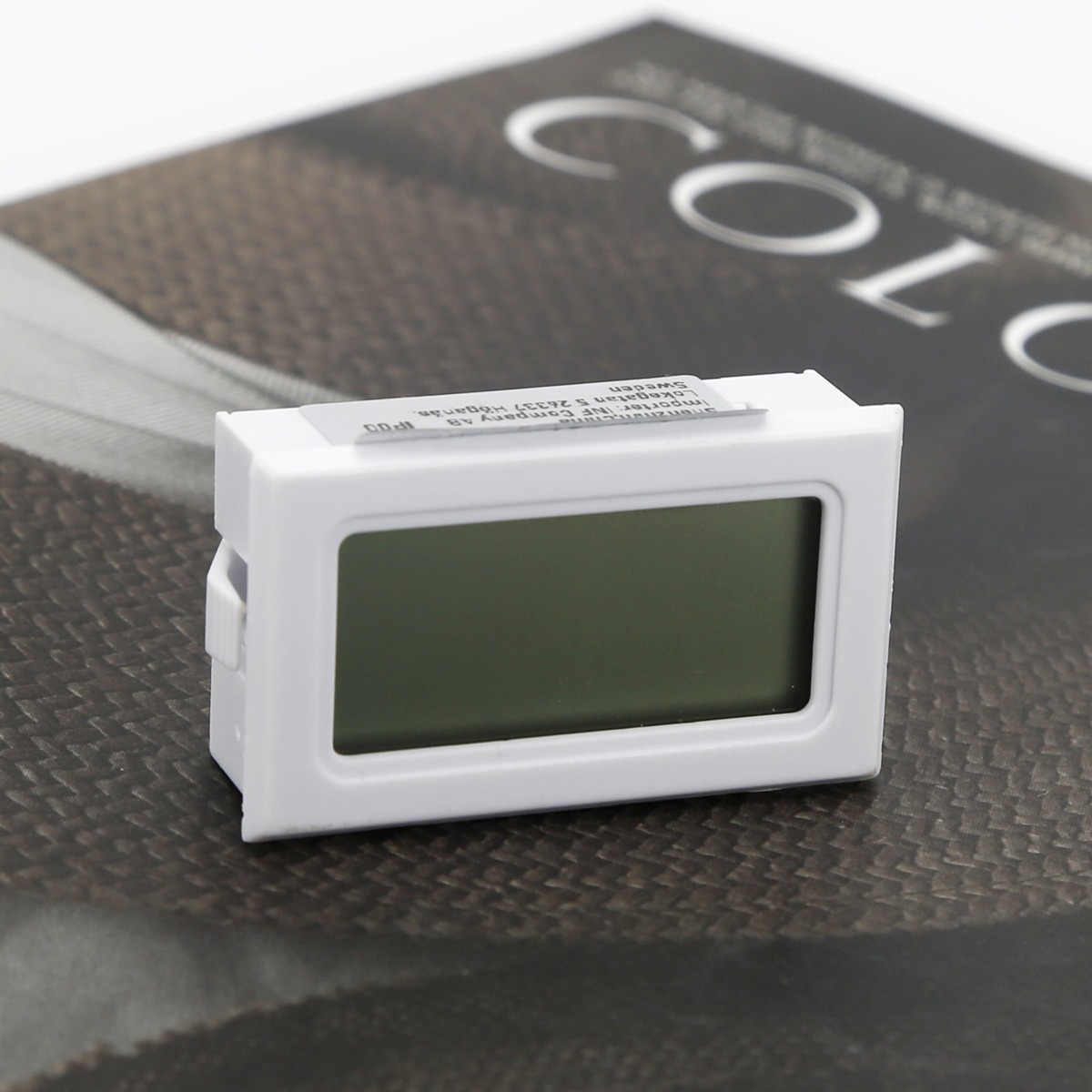 Ein kleines, weißes Digitalthermometer mit leerem Bildschirm liegt auf einer dunklen Musteroberfläche.
