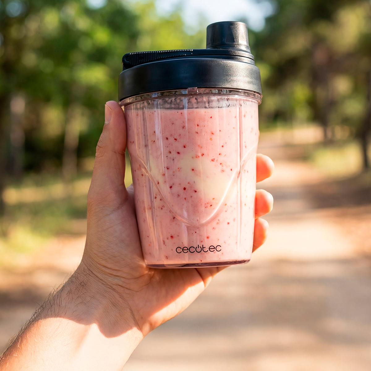 Una mano sostiene un vaso portátil lleno de un batido de frutas rosa al aire libre.