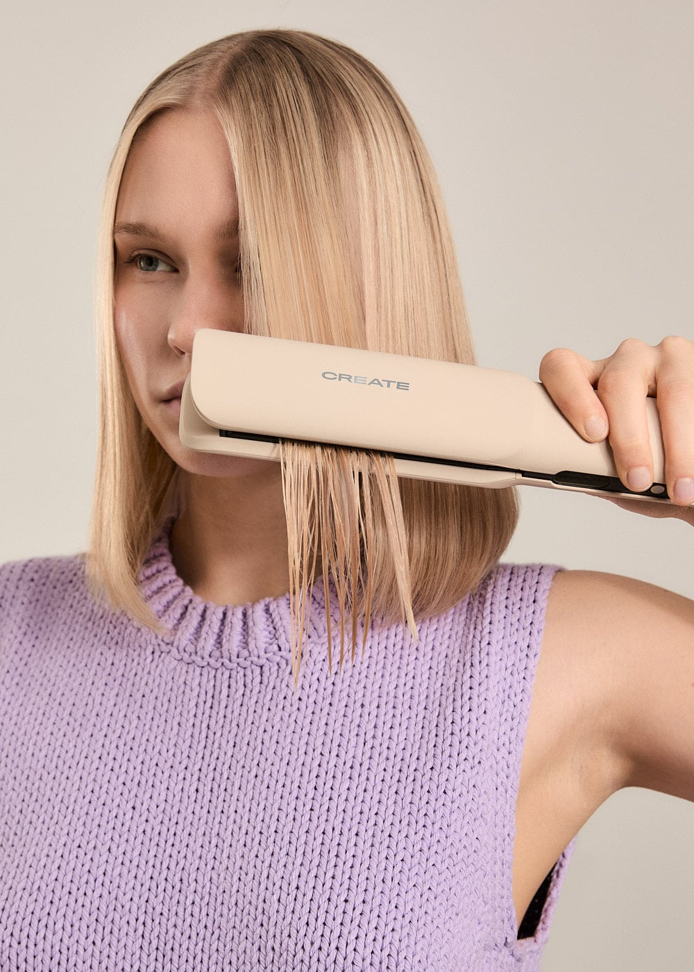 Una mujer rubia con un suéter de punto morado usa una plancha de pelo beige.
