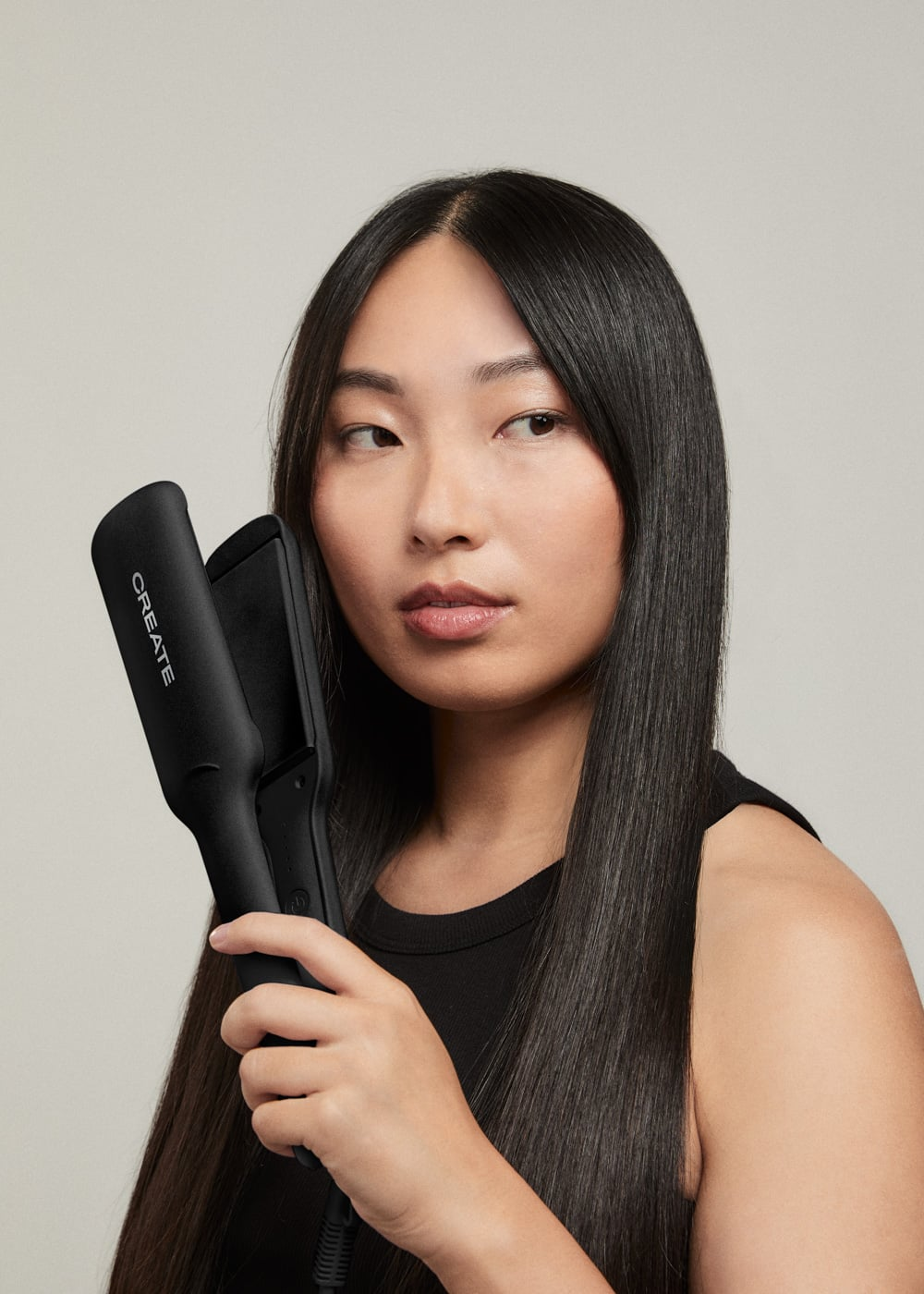 Una mujer con cabello negro largo sostiene una plancha de pelo negra sobre un fondo gris.