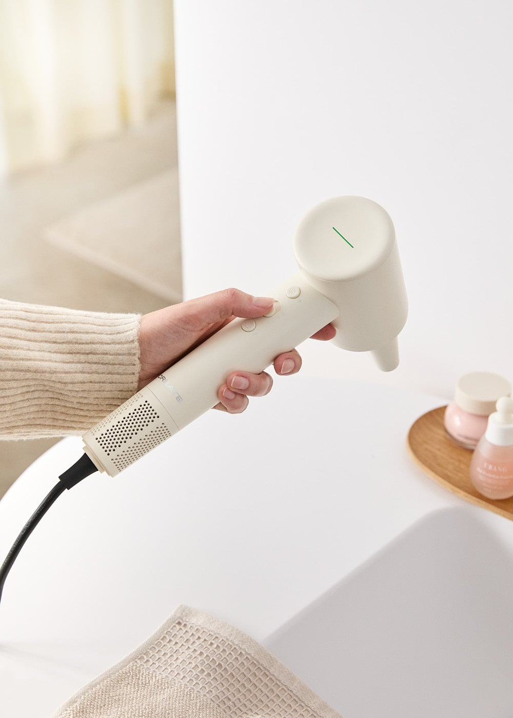Una mano sostiene un secador de pelo moderno y beige en un baño luminoso.