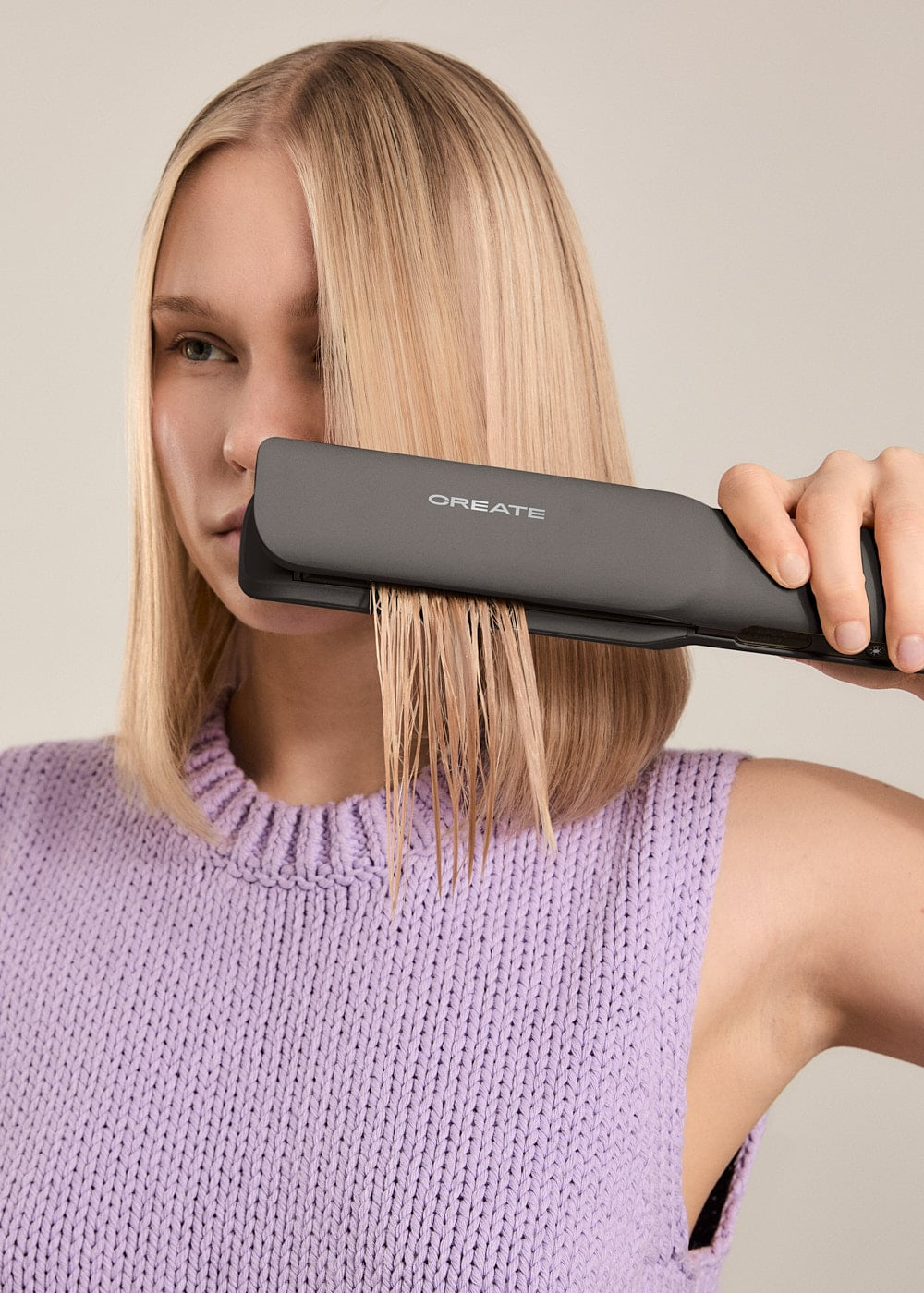 Una mujer rubia con top morado alisa su cabello mojado con una plancha gris.