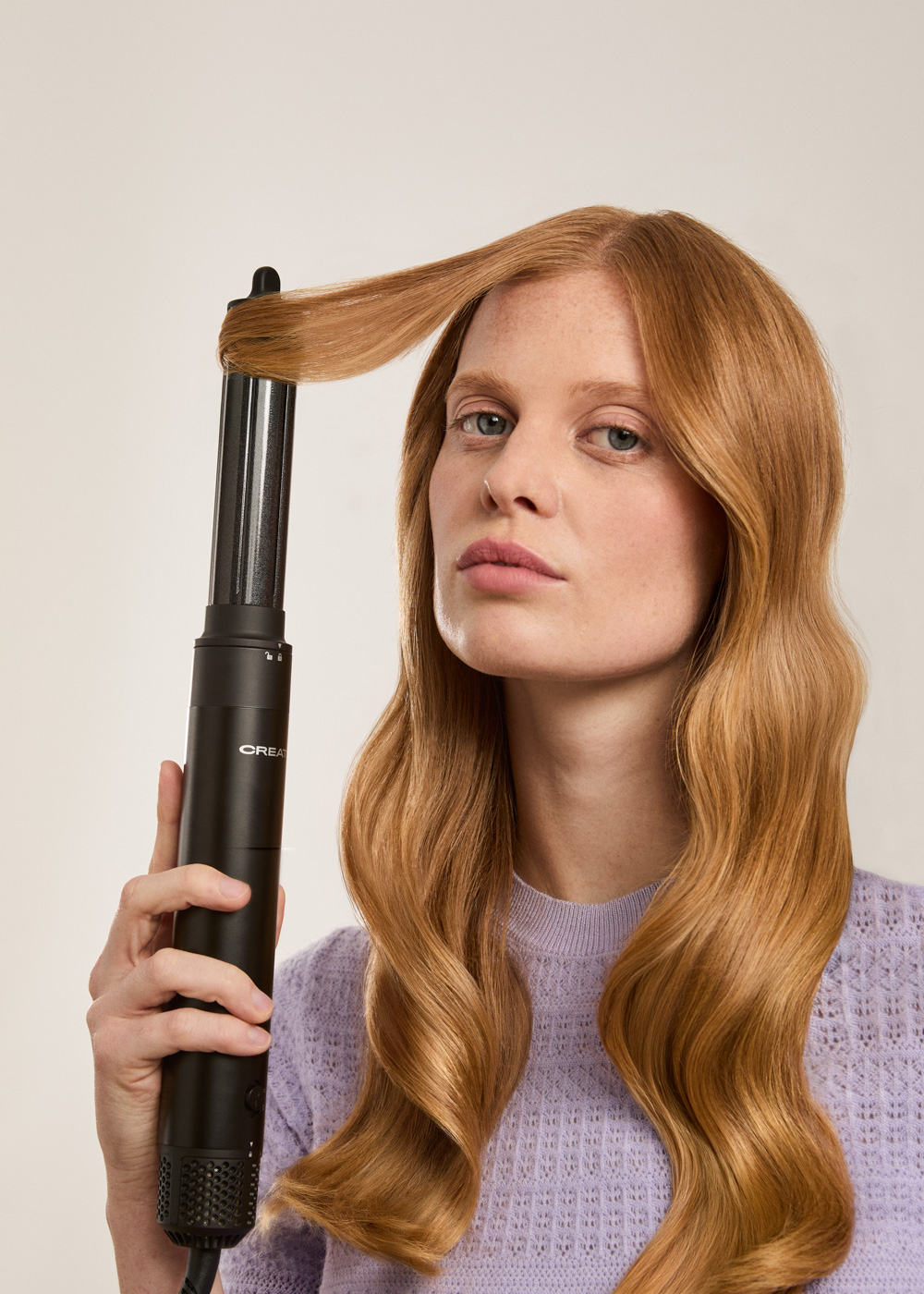 Una mujer de cabello rojo usa un rizador negro para peinar un mechón de cabello sobre fondo claro.