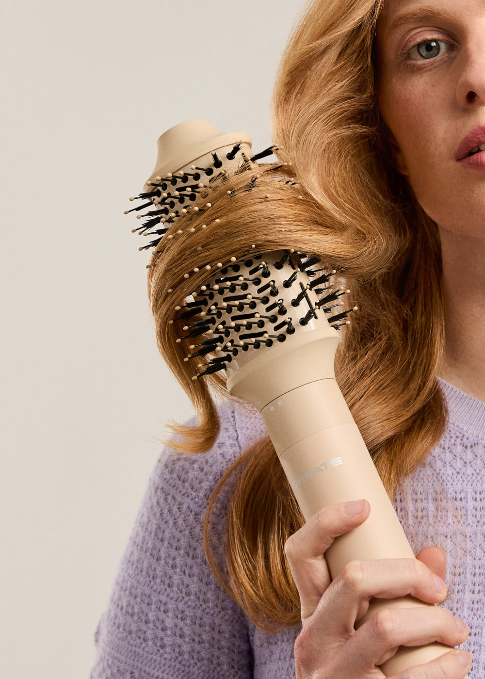 Una mujer sostiene un cepillo moldeador beis, peinando una sección de su cabello rubio ondulado sobre fondo liso.