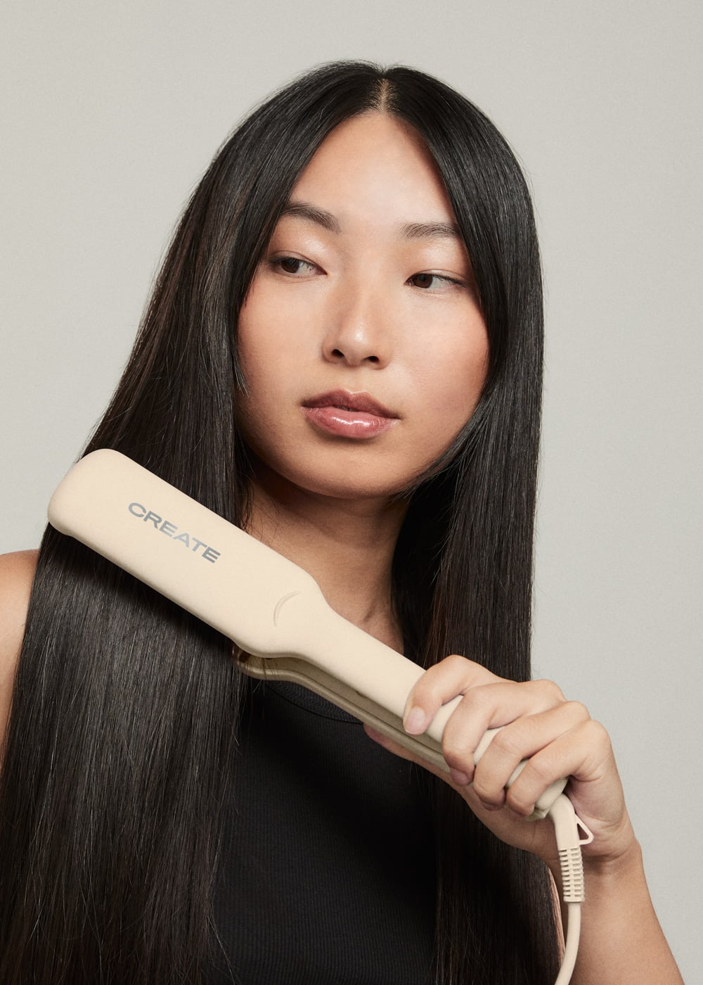 Una mujer con cabello negro, largo y lacio sostiene una plancha para el cabello beige contra un fondo gris.