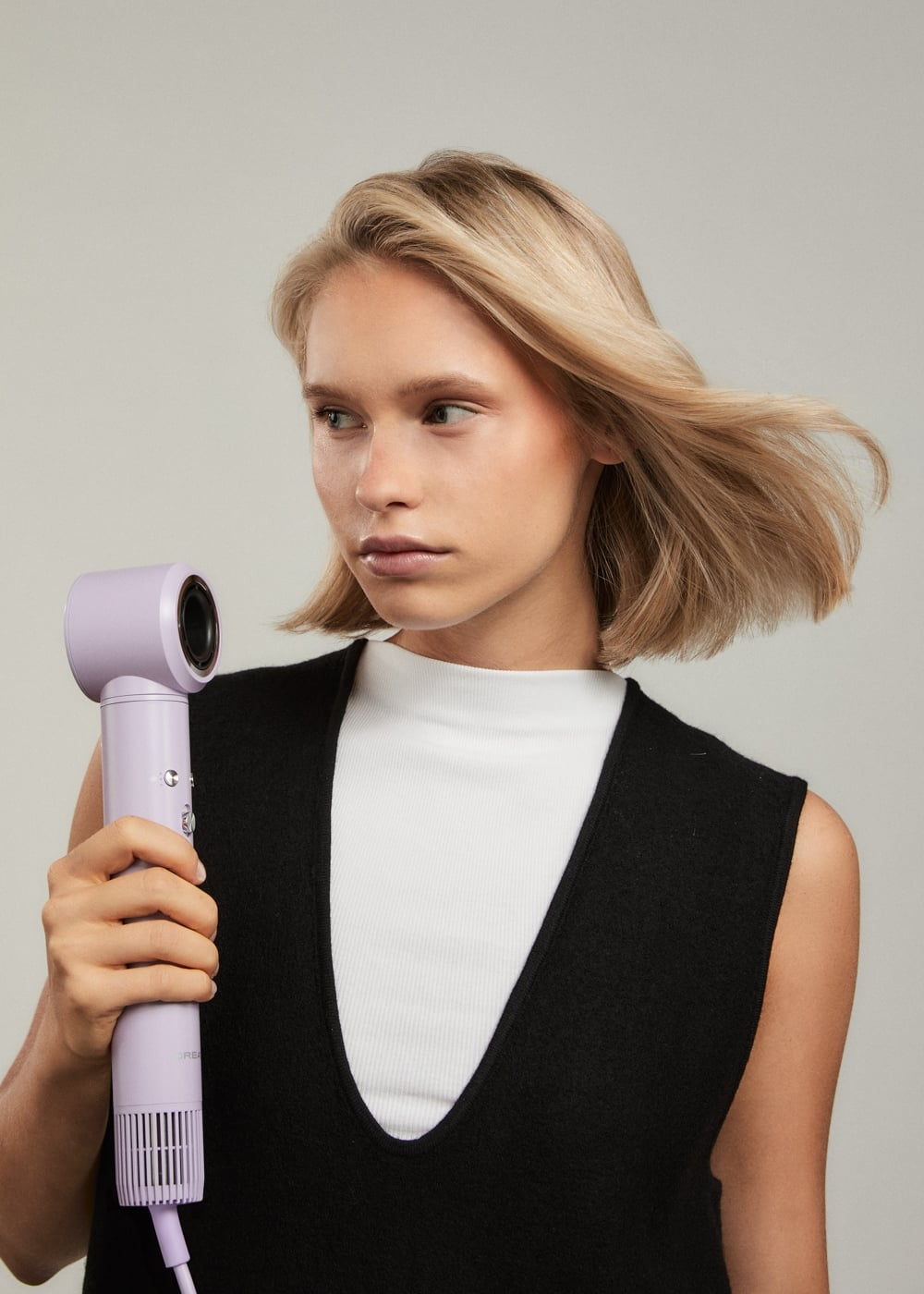 Una mujer rubia con top blanco y negro sostiene un secador morado mientras su cabello se mueve.