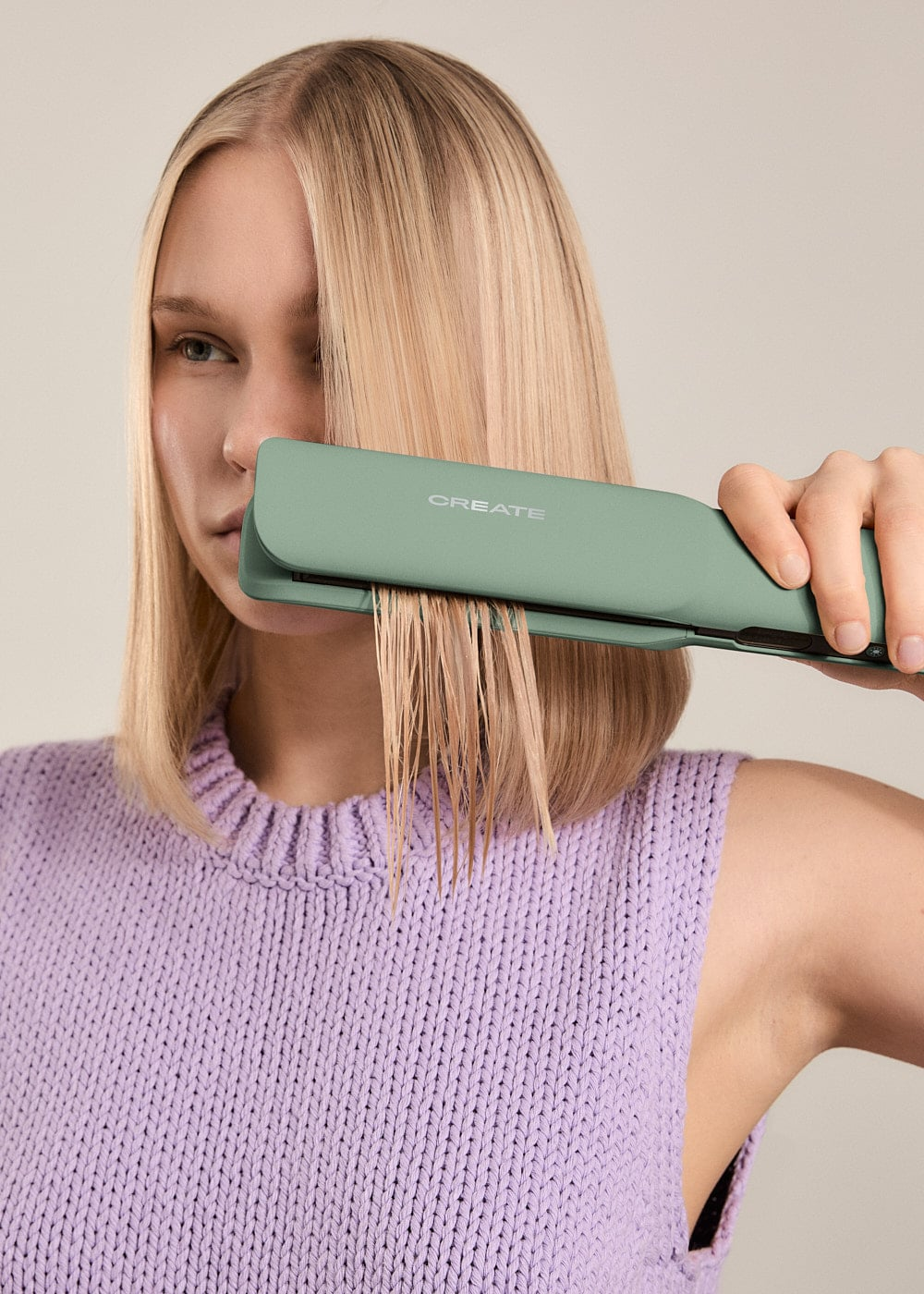 Una mujer rubia con top morado usa una plancha de pelo verde menta en cabello mojado.