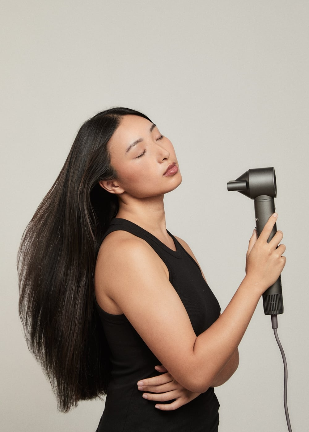 Una mujer con cabello negro largo sostiene un secador de pelo gris sobre un fondo claro.