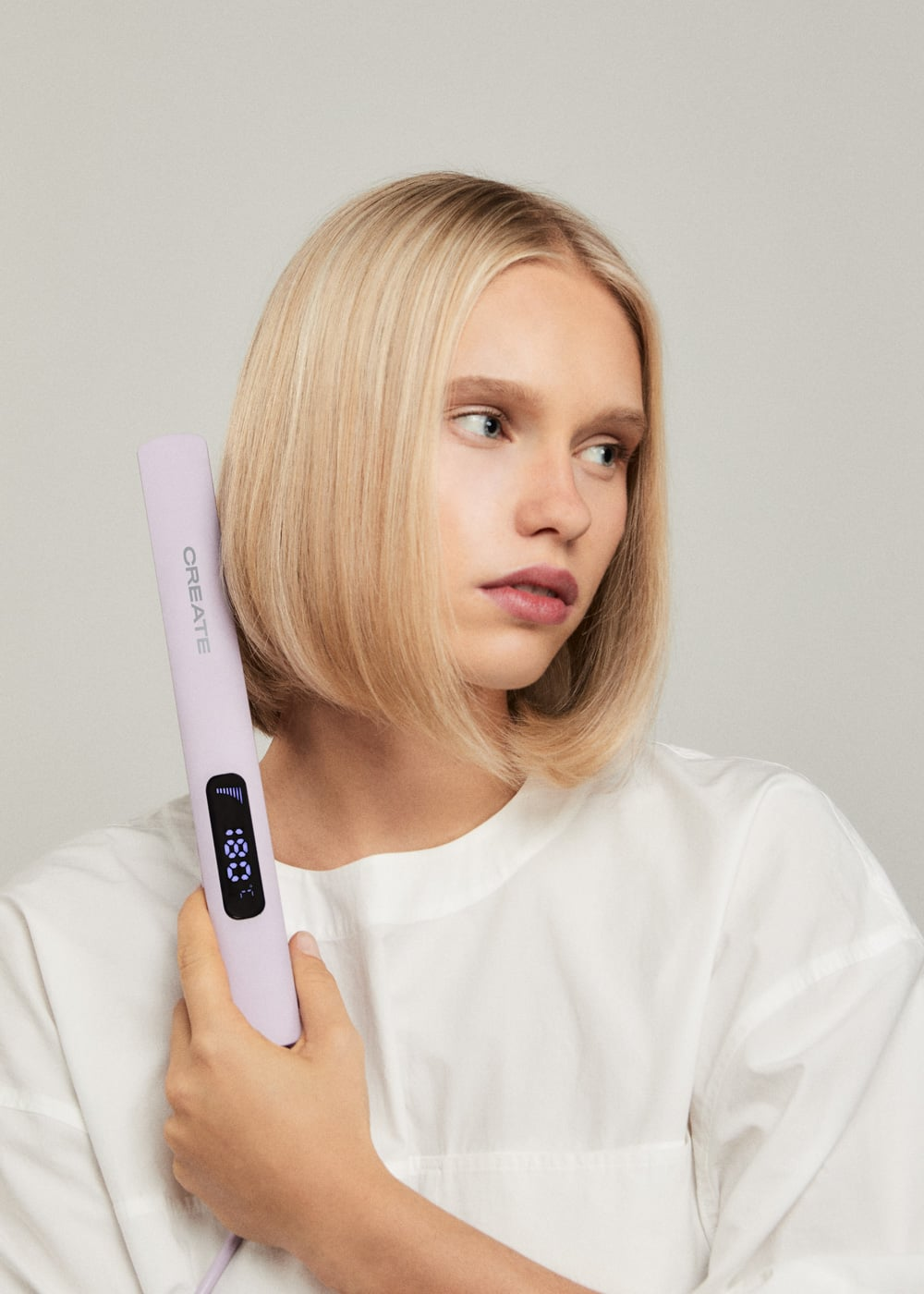 Una mujer rubia con camisa blanca sostiene una plancha de pelo lavanda sobre un fondo neutro.