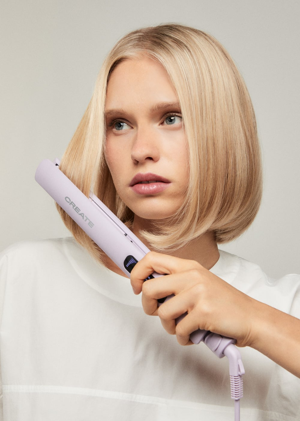 Una mujer rubia usa una plancha de pelo morada clara, moldeando su pelo corto sobre un fondo gris neutro.