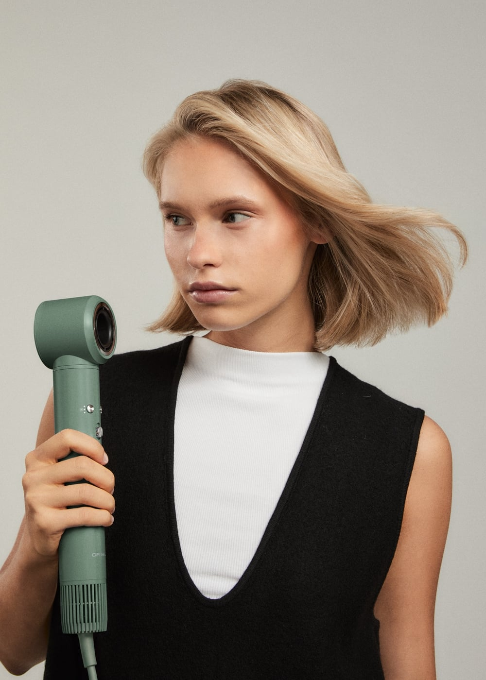 Una mujer rubia con chaleco negro sostiene un secador verde menta, con el cabello al viento.