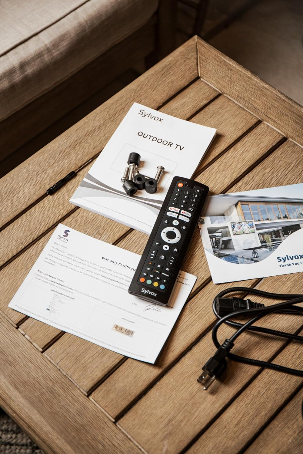 Un manual de TV para exteriores, certificado de garantía, control remoto y cable de alimentación en una mesa.