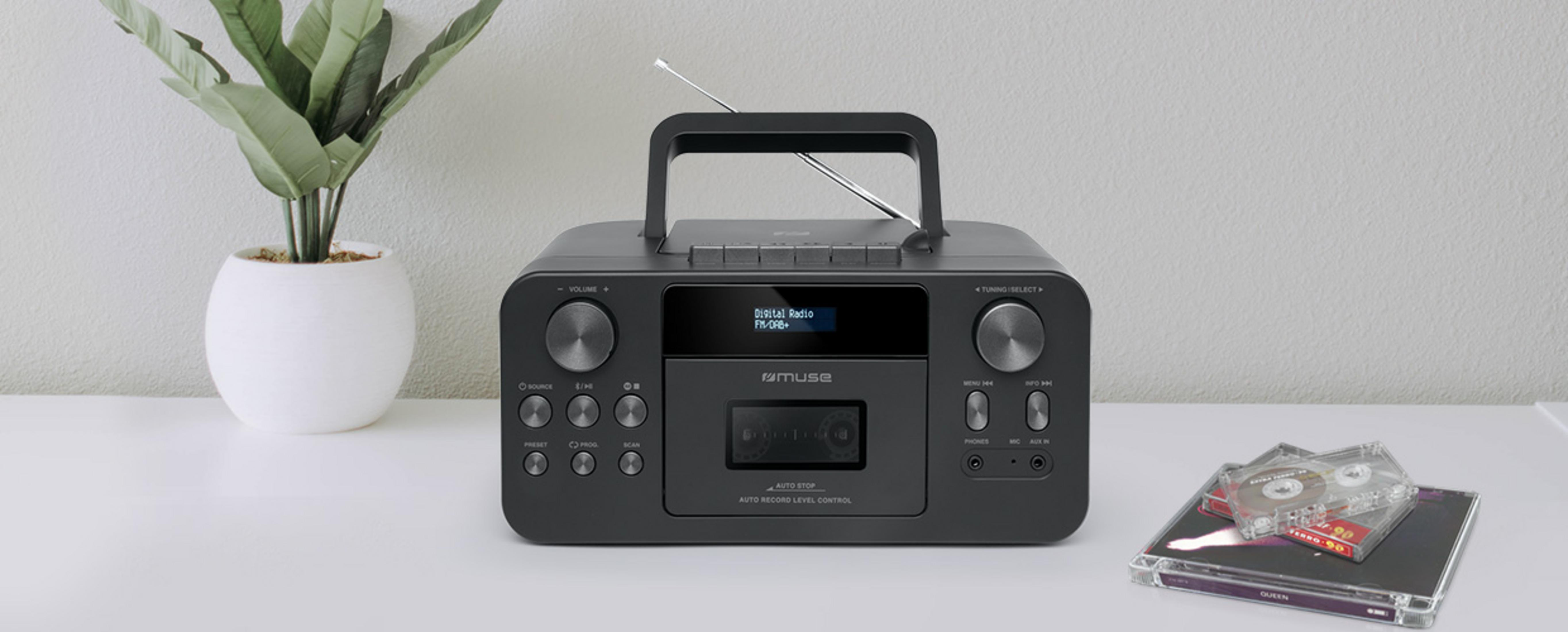Ein schwarzes Kofferradio auf weißem Tisch, links eine Pflanze im Topf und rechts Musikkassetten.