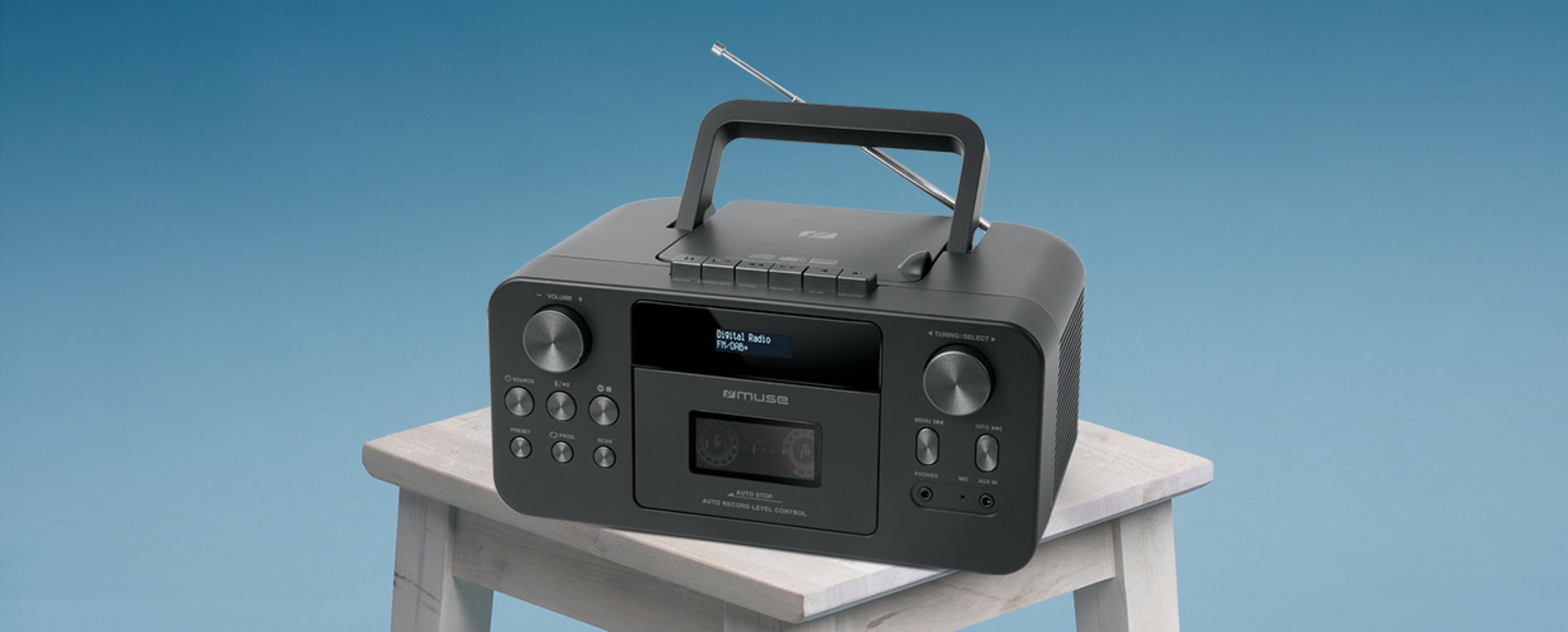 Ein schwarzes Kofferradio mit Kassettenlaufwerk und Antenne steht auf einem weißen Holzhocker vor blauem Hintergrund.