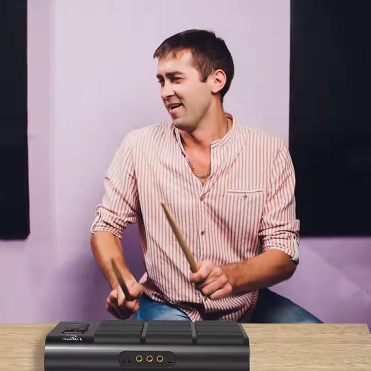 Ein Mann im gestreiften Hemd spielt an einem Holztisch mit Drumsticks auf einem E-Drum-Pad.
