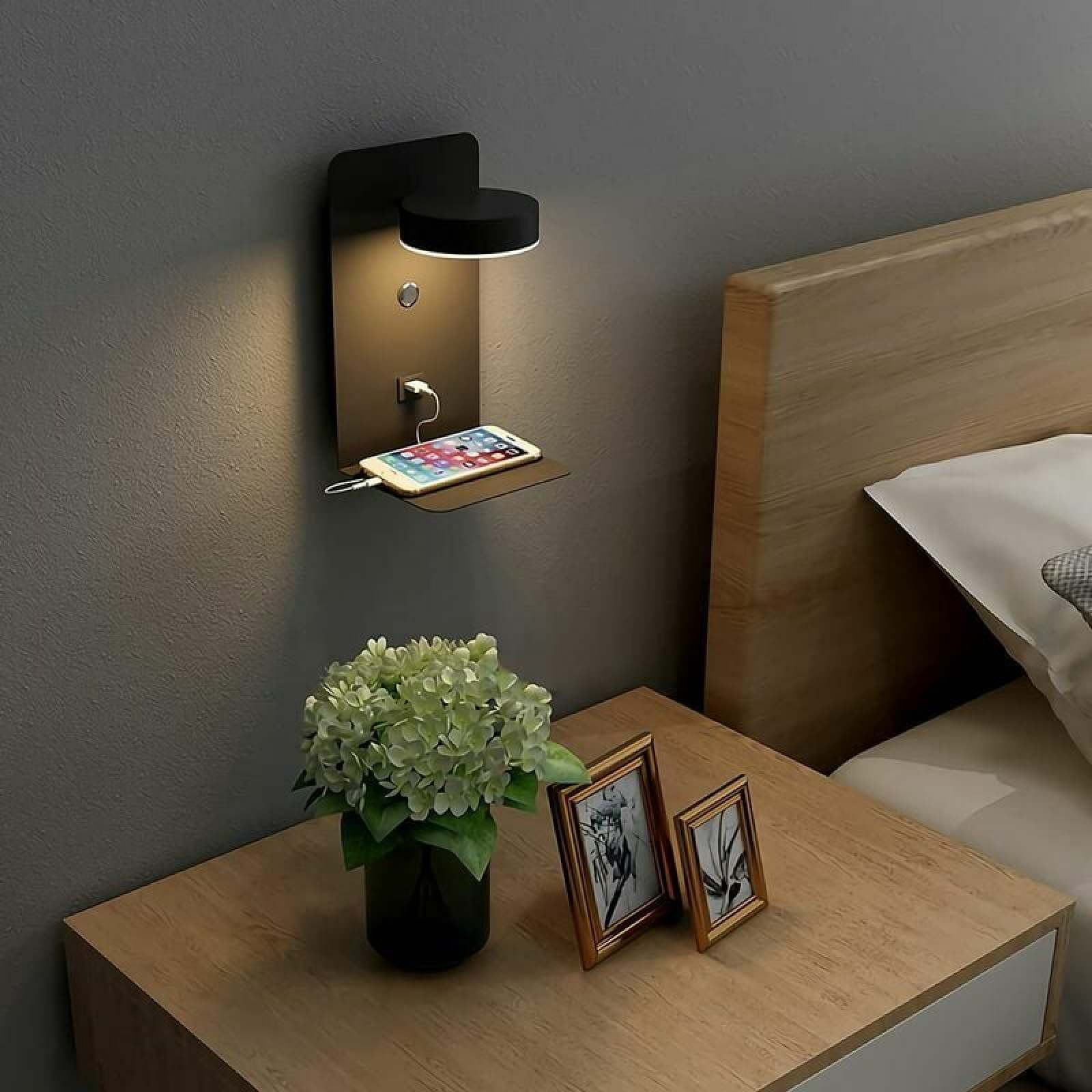 Schwarze Wandlampe mit Ladefunktion über dem Nachttisch, darauf Blumen und Fotorahmen neben dem Bett.