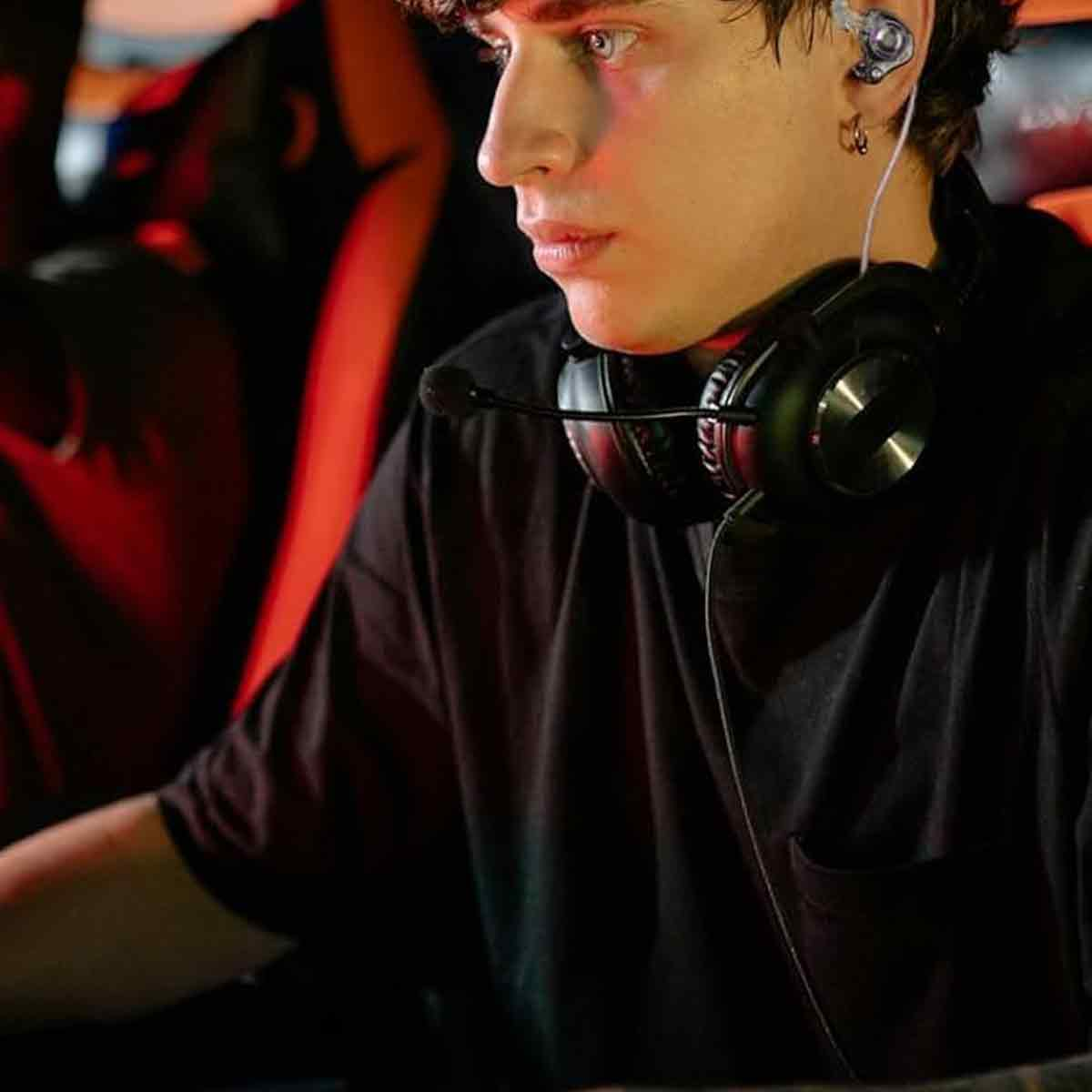 Ein junger Mann im schwarzen T-Shirt trägt ein Headset mit Mikrofon und sitzt vor einem roten Hintergrund.
