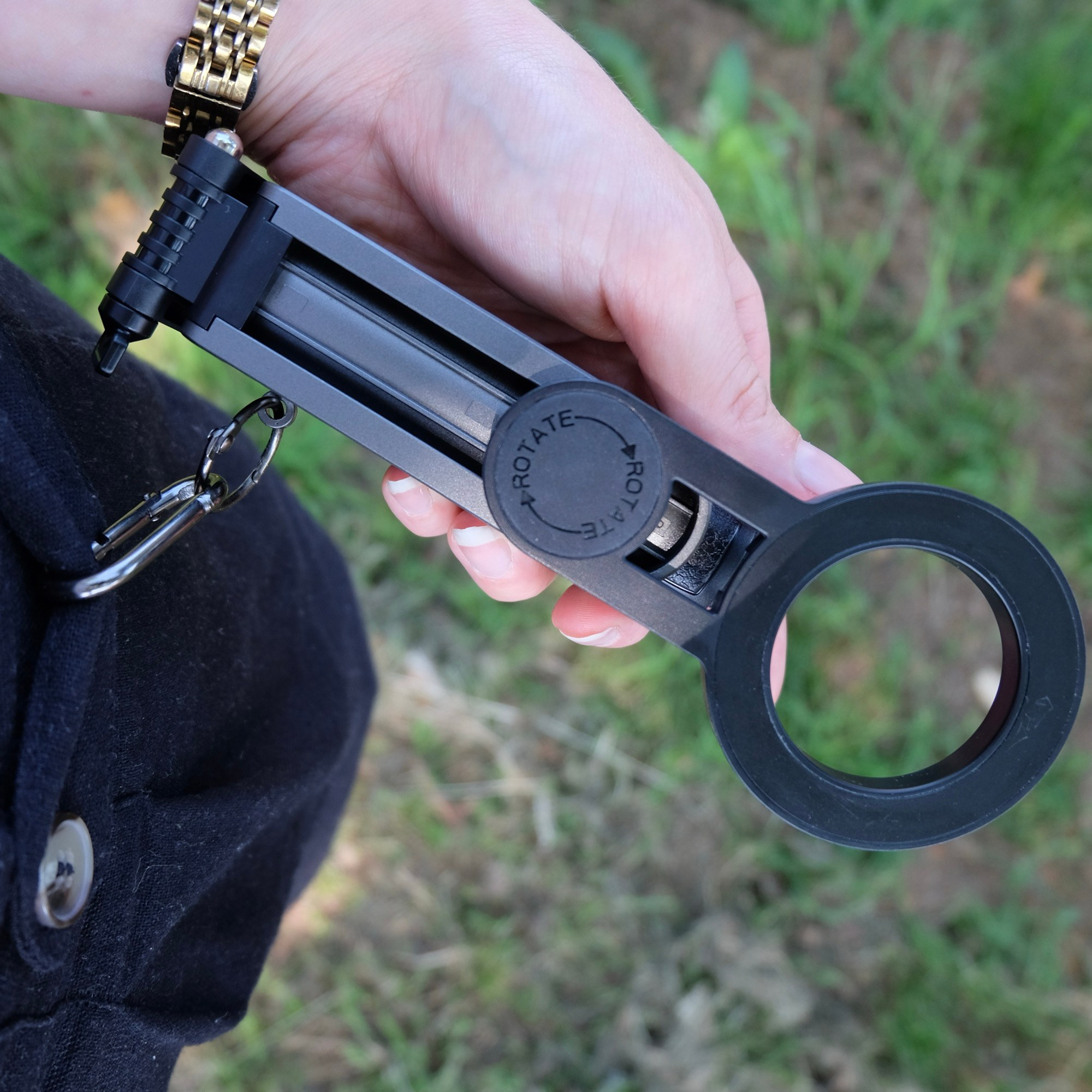 Eine Hand hält ein schwarzes, faltbares Gadget mit Ring und Aufschrift "Rotate" über grünem Gras.