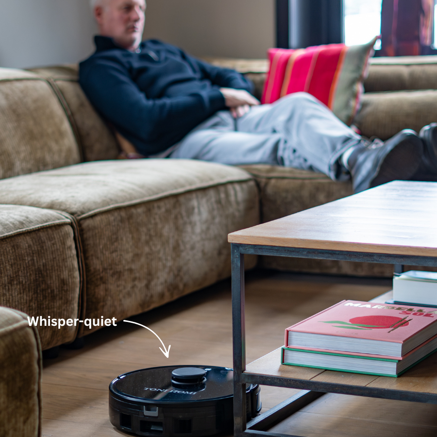 Ein schwarzer Saugroboter reinigt den Boden in einem Wohnzimmer, während ein Mann auf einem Sofa ausruht.