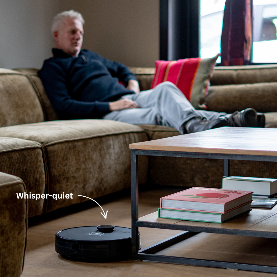 Ein schwarzer Saugroboter reinigt den Boden in einem Wohnzimmer, während ein Mann auf einem Sofa entspannt.