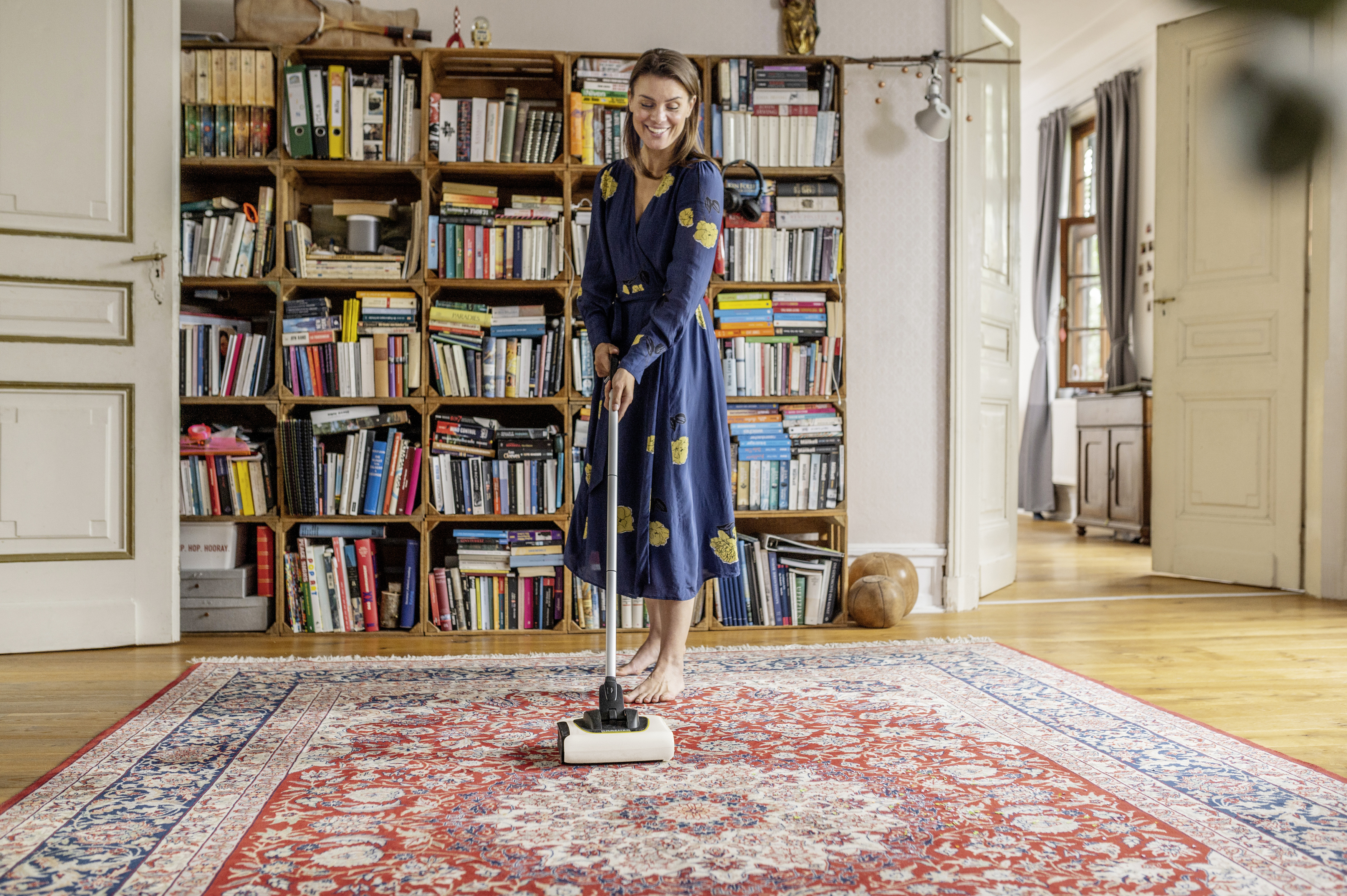 Una donna in abito blu passa l'aspirapolvere su un tappeto rosso davanti a una libreria.