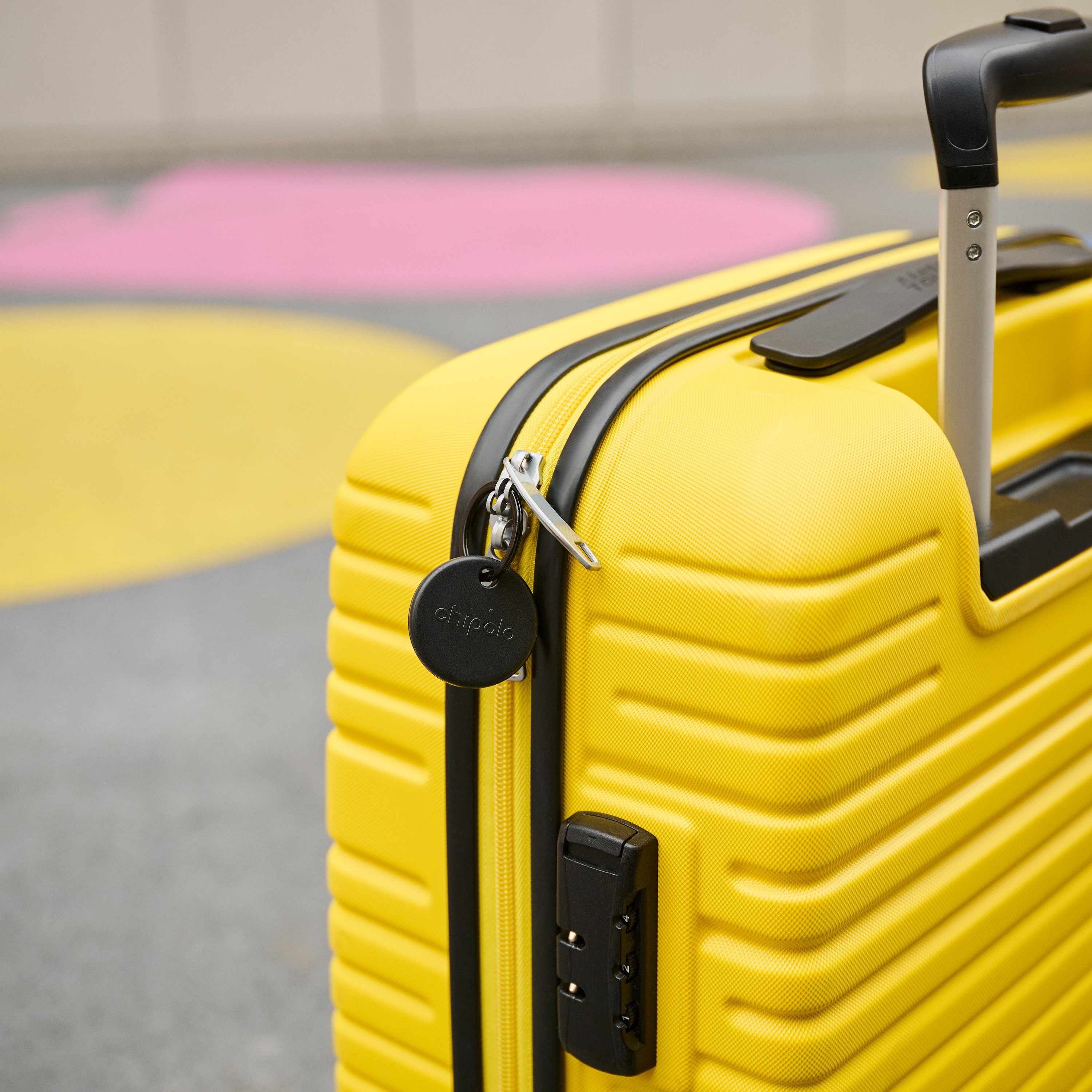 Une valise jaune avec un traceur Chipolo noir attaché à la fermeture sur un sol coloré.