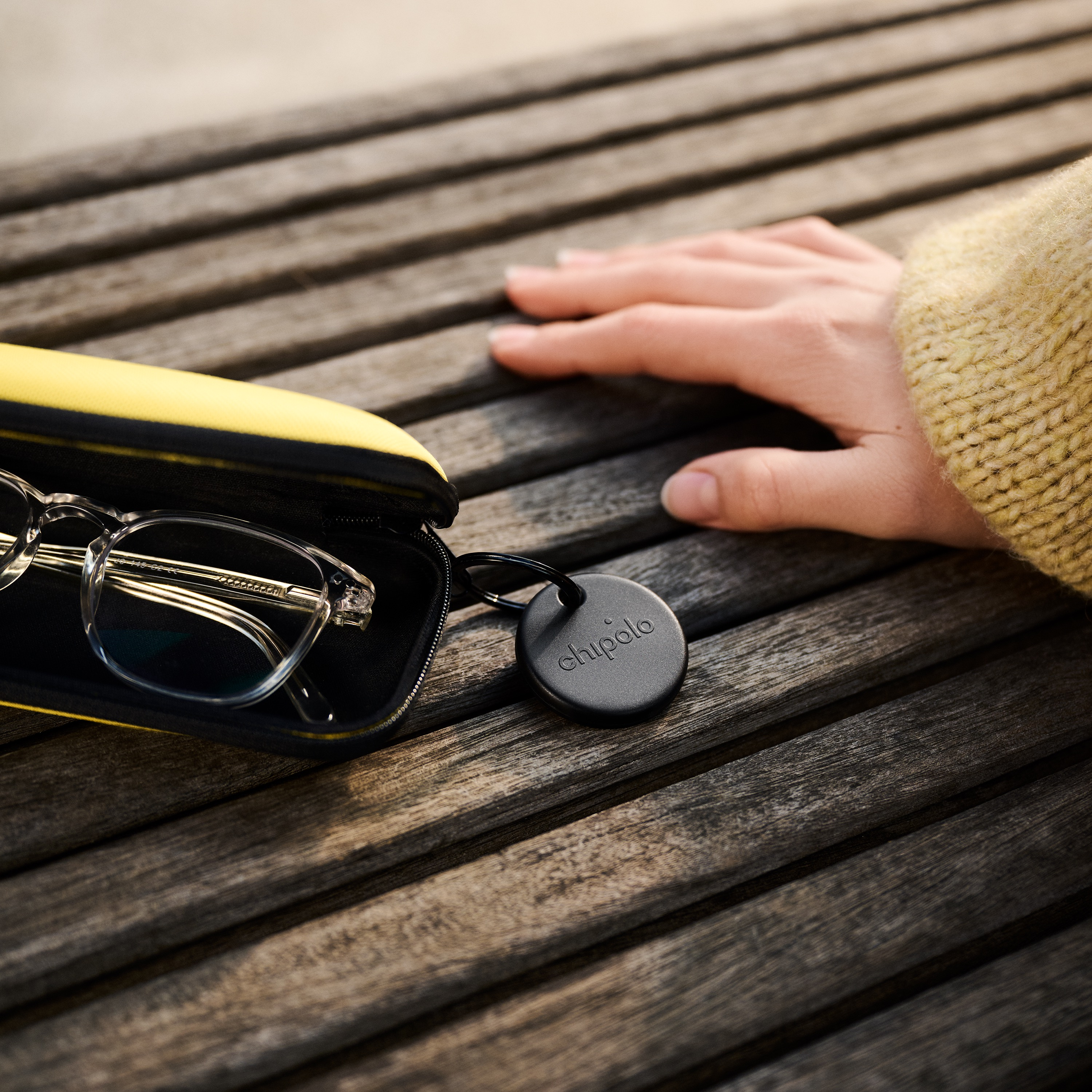 Une main repose sur un banc en bois à côté d'un étui à lunettes ouvert contenant des lunettes transparentes et un traceur Chipolo noir.