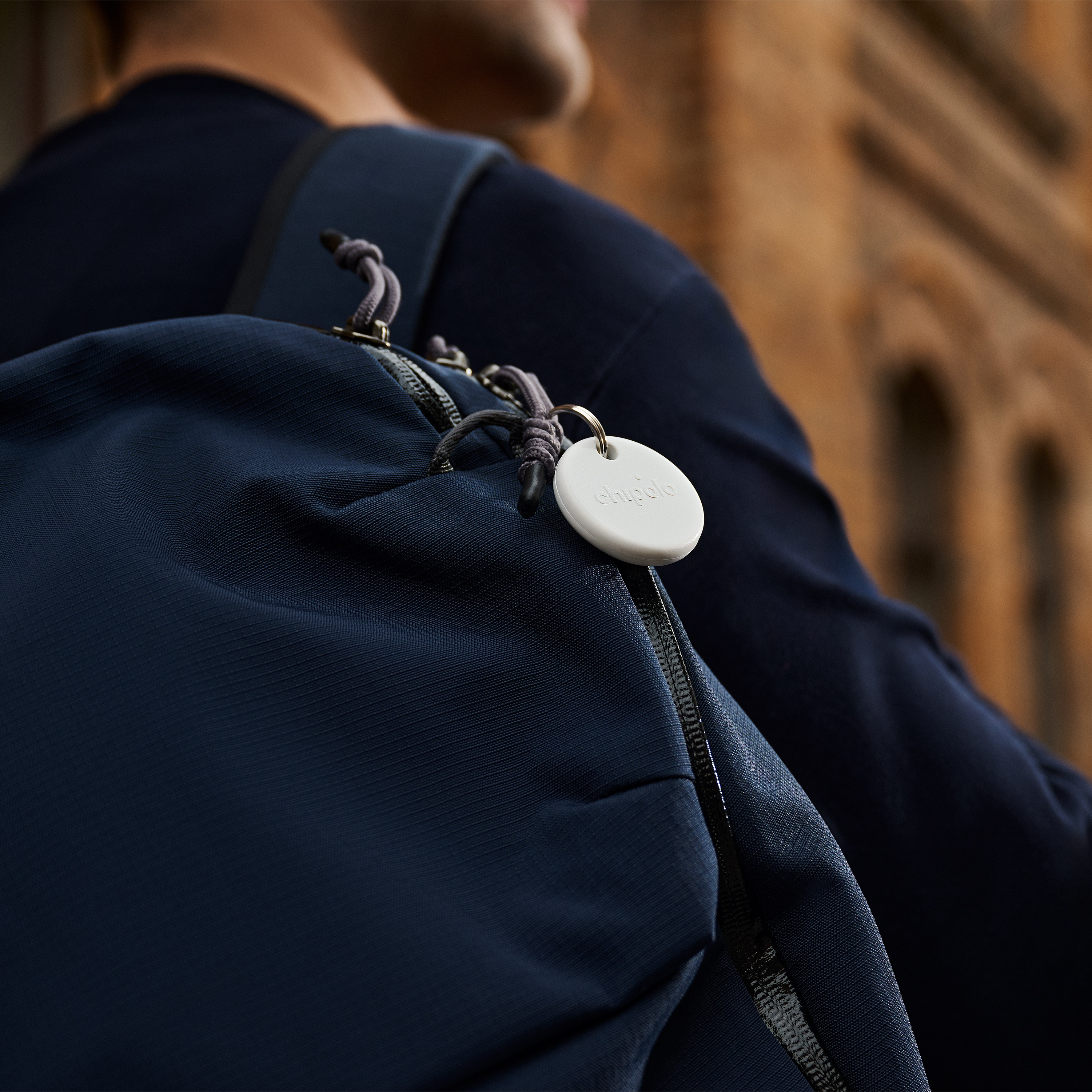 Un traceur Chipolo blanc est suspendu à la fermeture éclair d'un sac à dos bleu porté par une personne.