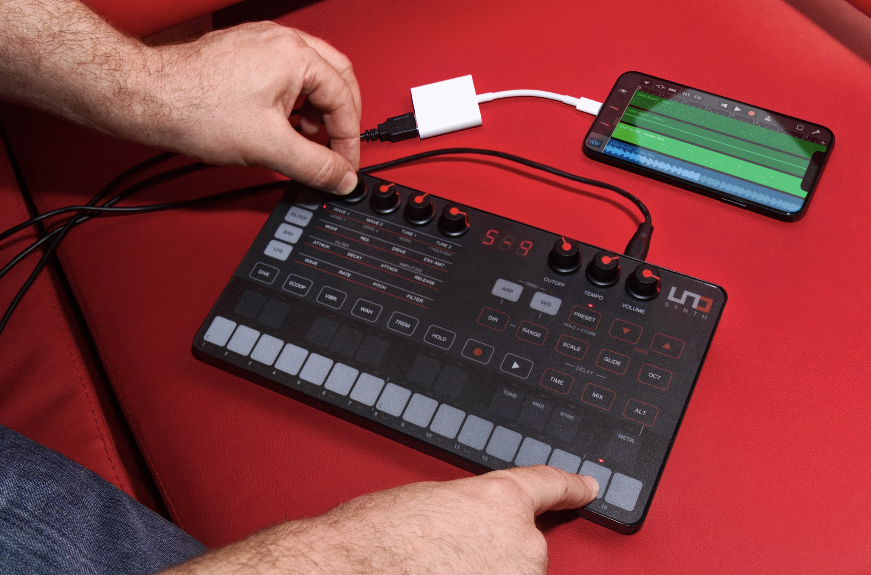 Eine Hand dreht einen Regler am schwarzen Uno Synth auf rotem Hintergrund, daneben ein Smartphone mit Musik-App.