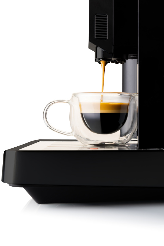 Eine schwarze Kaffeemaschine gießt frischen Espresso in eine doppelwandige Glastasse vor weißem Hintergrund.