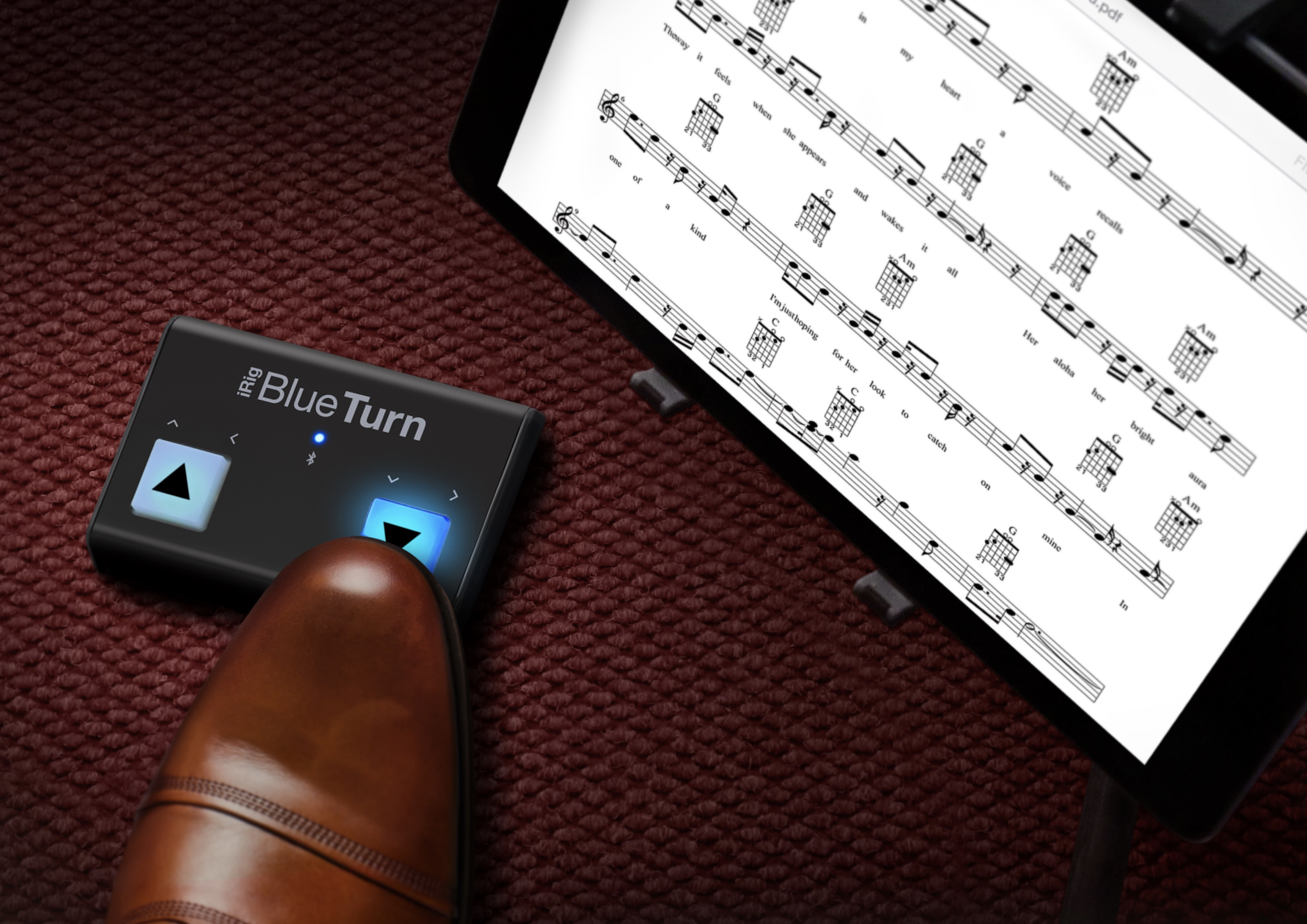 Ein brauner Lederschuh drückt ein BlueTurn-Pedal neben einem Noten-Tablet auf rotem Teppich.