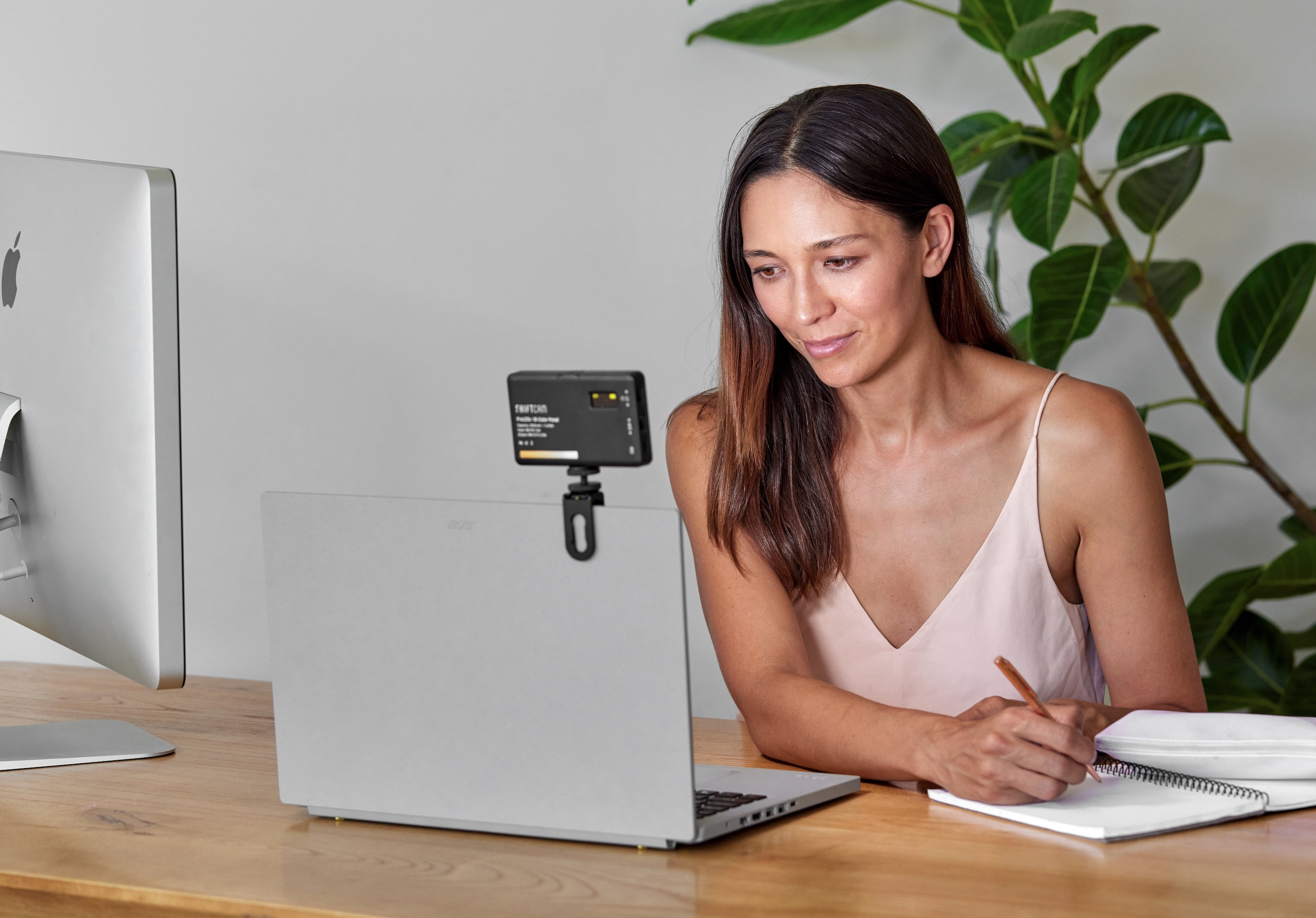Eine Frau sitzt am Holztisch vor einem Laptop mit angeclipster Lichtquelle und schreibt in ein Notizbuch.