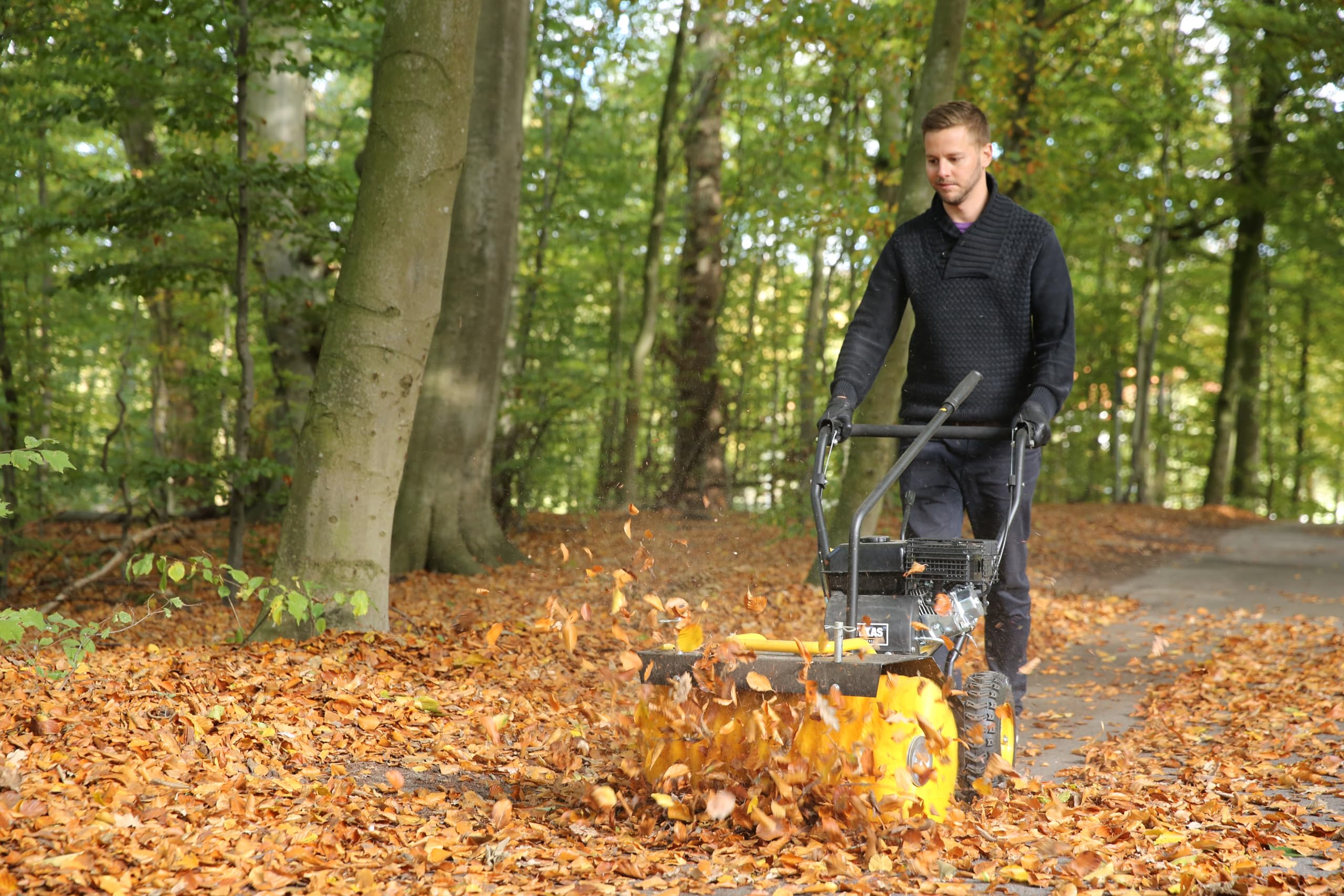 Ein Mann im schwarzen Pullover nutzt einen Laubsauger auf einem herbstlichen Waldweg.
