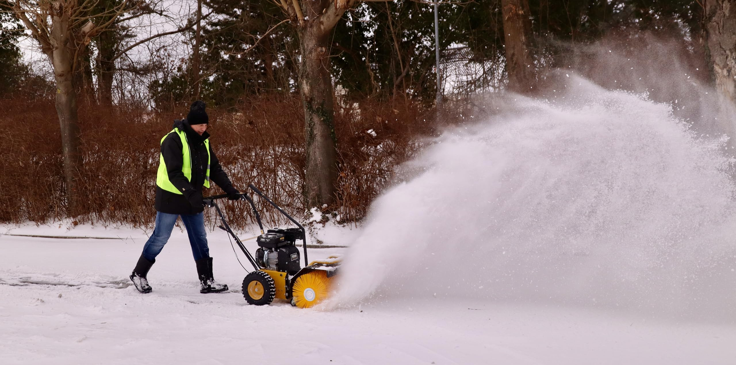 Eine Person in gelber Weste bedient im winterlichen Park eine gelbe Kehrmaschine, die Schnee aufwirbelt.