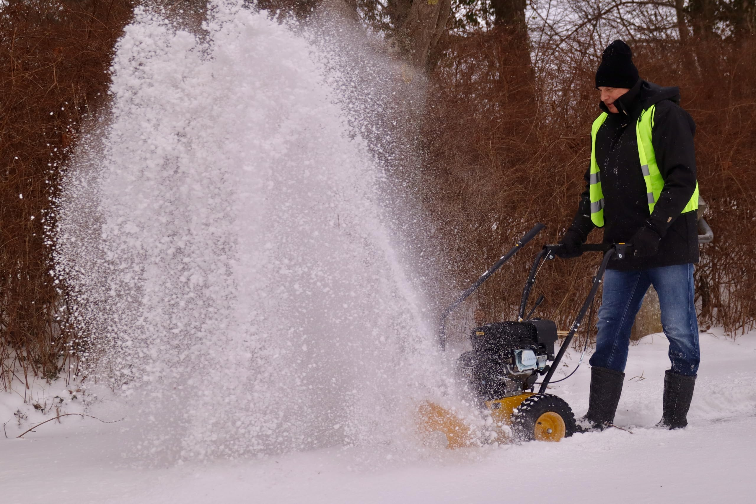 Ein Mann im schwarzen Wintermantel und gelber Warnweste benutzt eine Schneefräse vor kahlen Büschen.