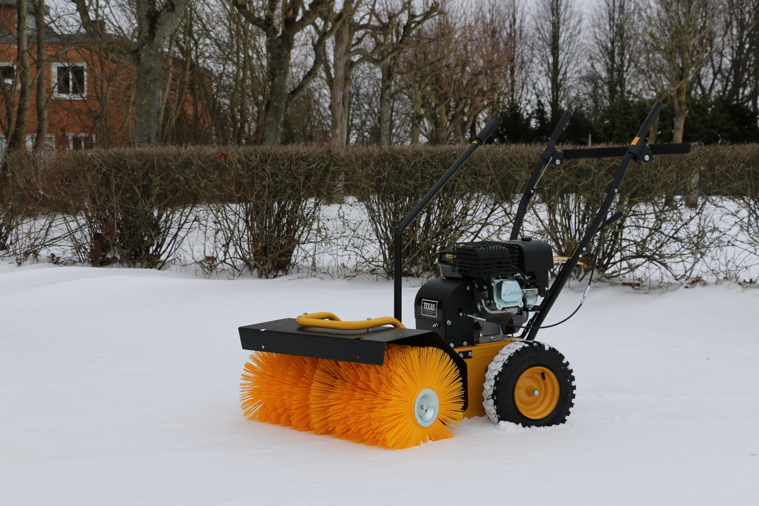 Eine gelbe und schwarze Motorbürste steht im Schnee vor einer Hecke und Bäumen.
