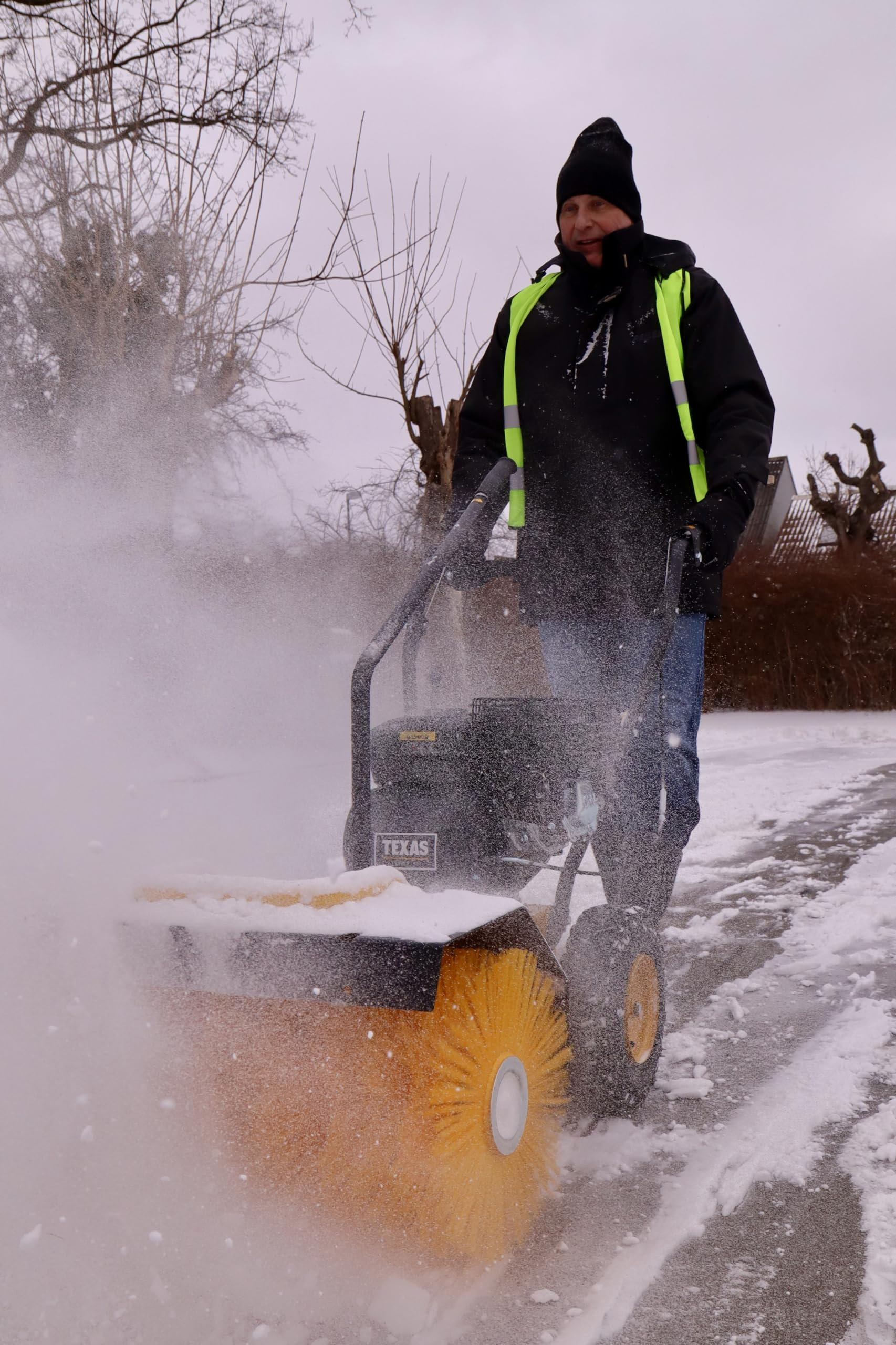 Ein Mann in schwarzer Jacke und Warnweste bedient eine gelbe rotierende Schneebürste auf einem schneebedeckten Weg.