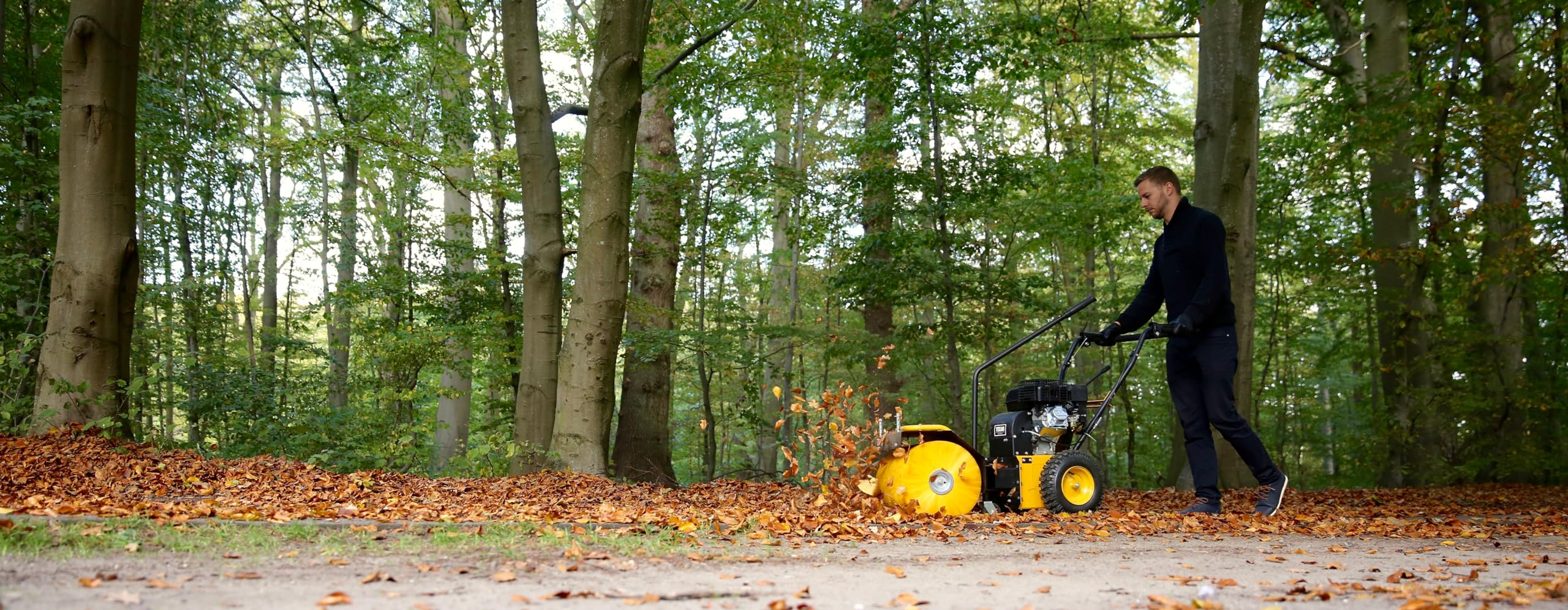 Ein Mann nutzt eine gelbe Kehrmaschine, um Laub auf einem Waldweg zu räumen.