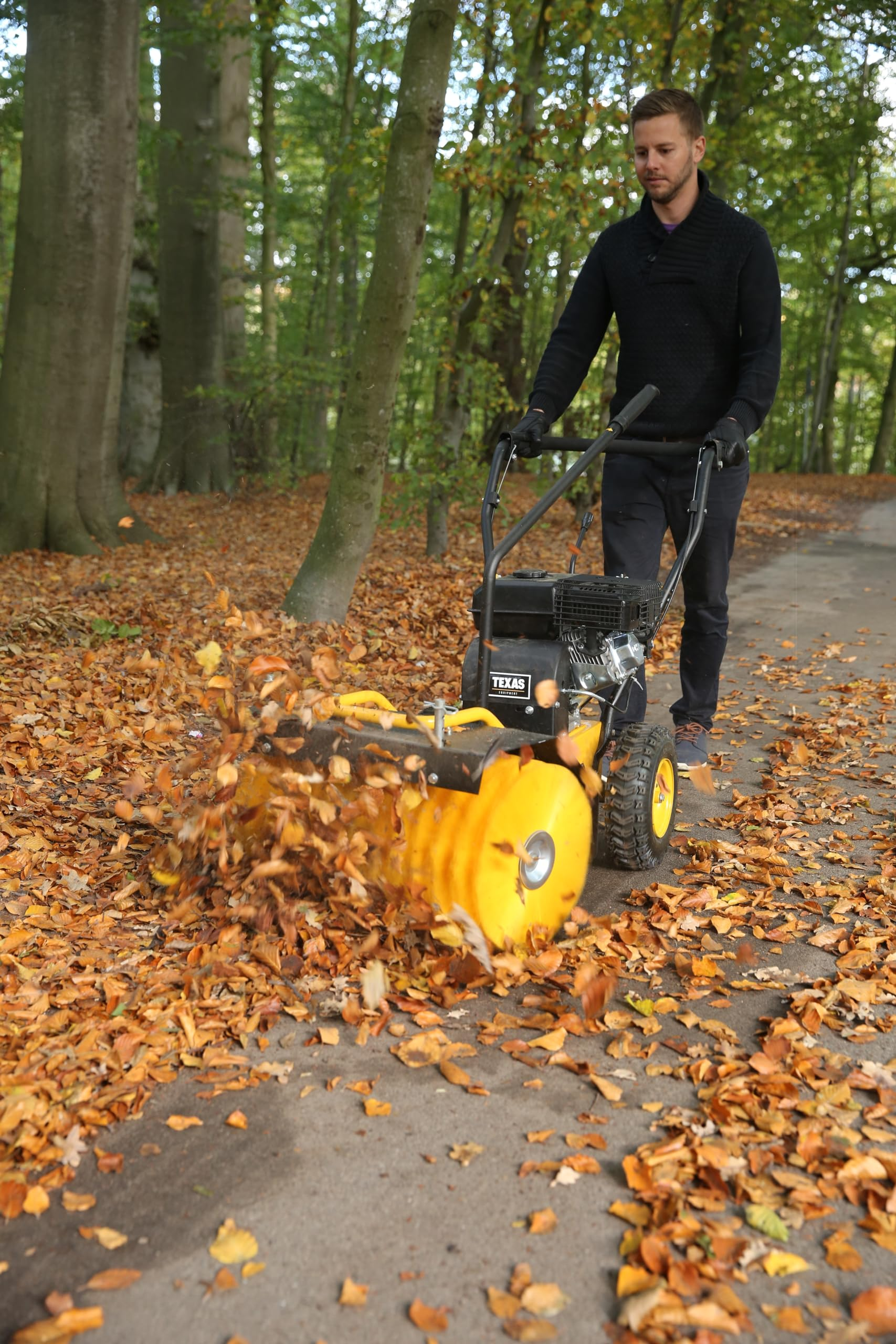 Ein Mann bedient eine gelbe Kehrmaschine und räumt Herbstblätter auf einem Parkweg.