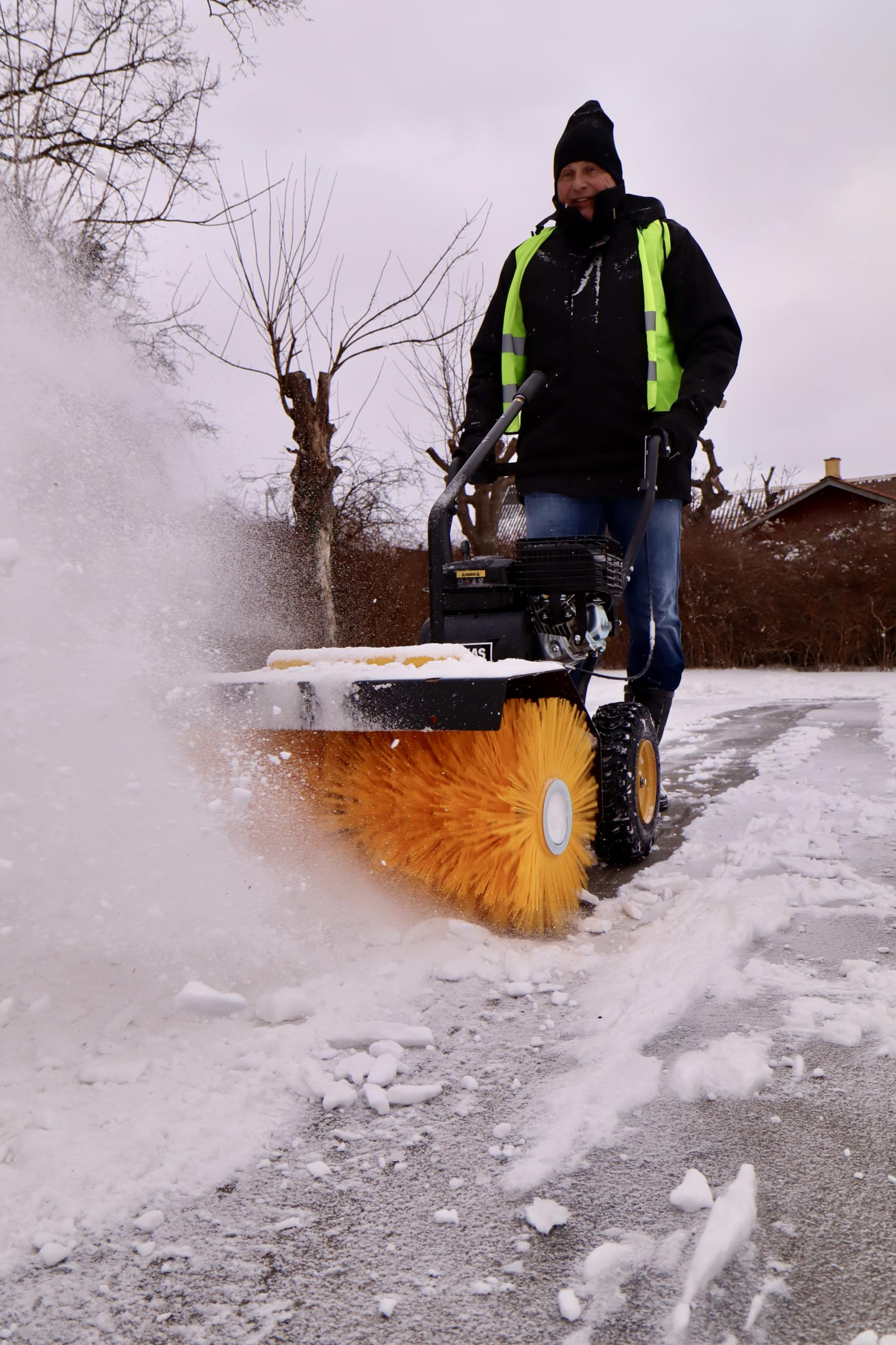 Ein Mann in Warnweste bedient im Winter eine gelbe Motorbürste auf schneebedecktem Boden.