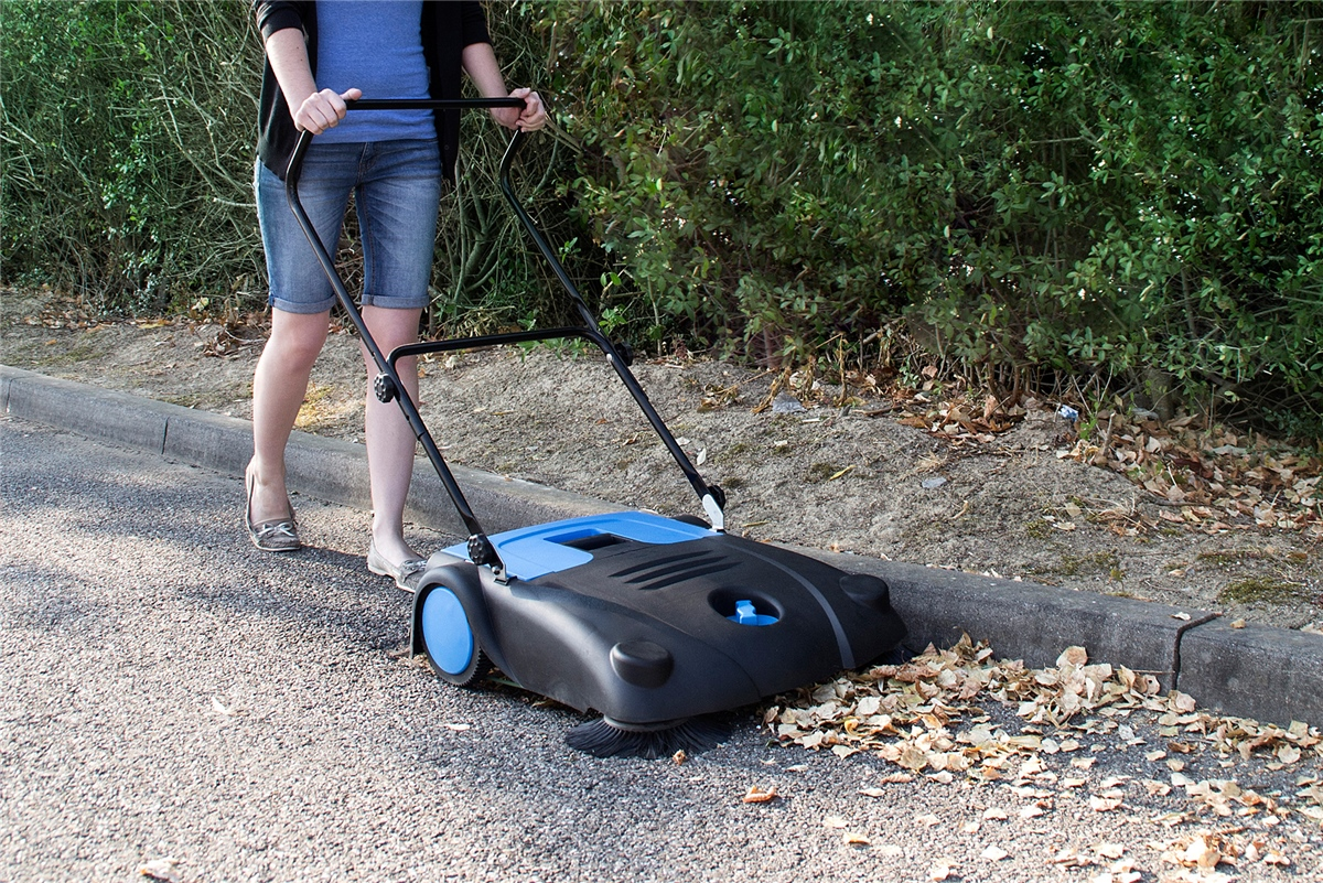 Eine Frau schiebt eine blaue und schwarze Kehrmaschine auf einem asphaltierten Weg.