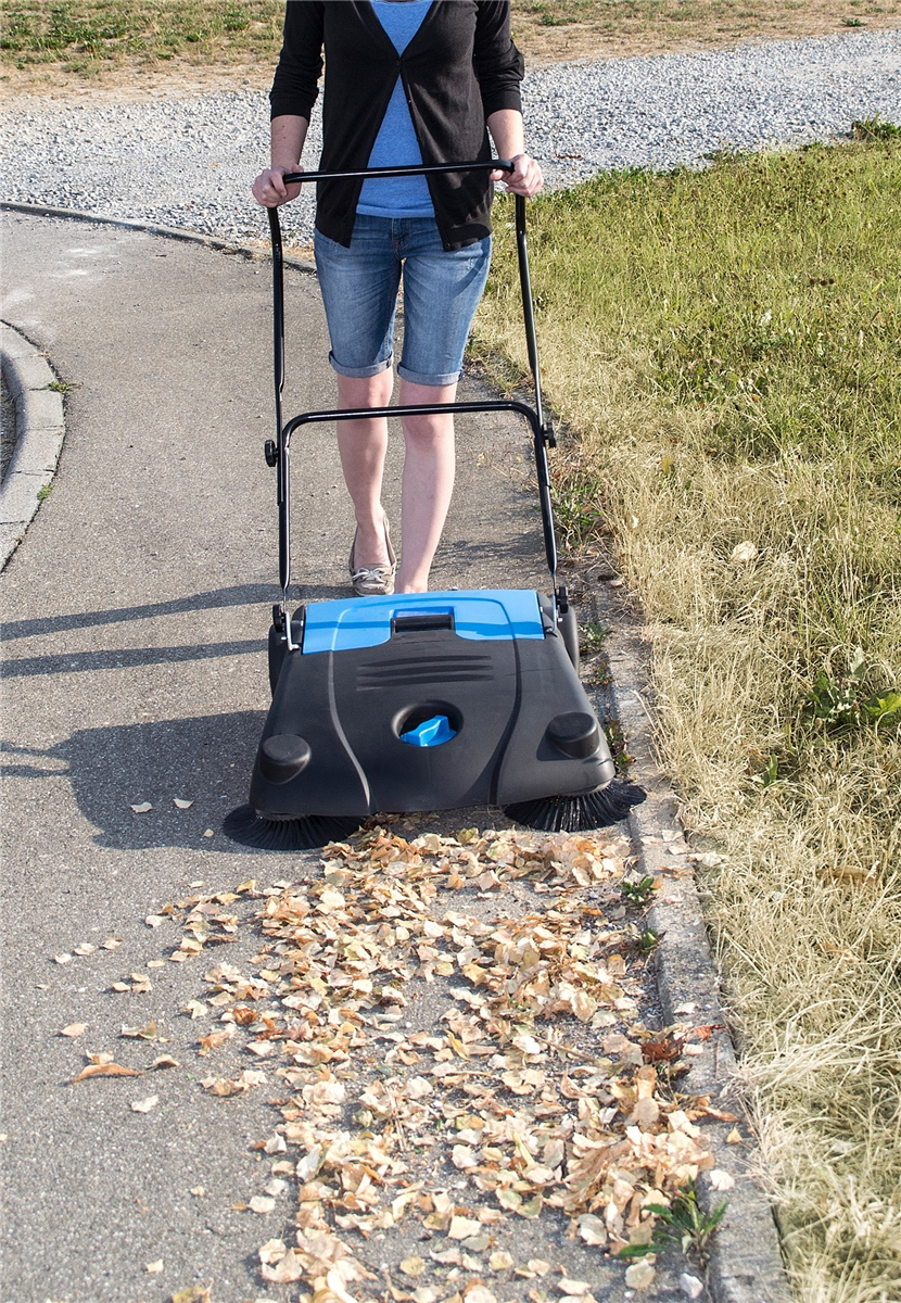 Eine Person schiebt eine blaue und schwarze Kehrmaschine auf einem Gehweg und sammelt Laub auf.