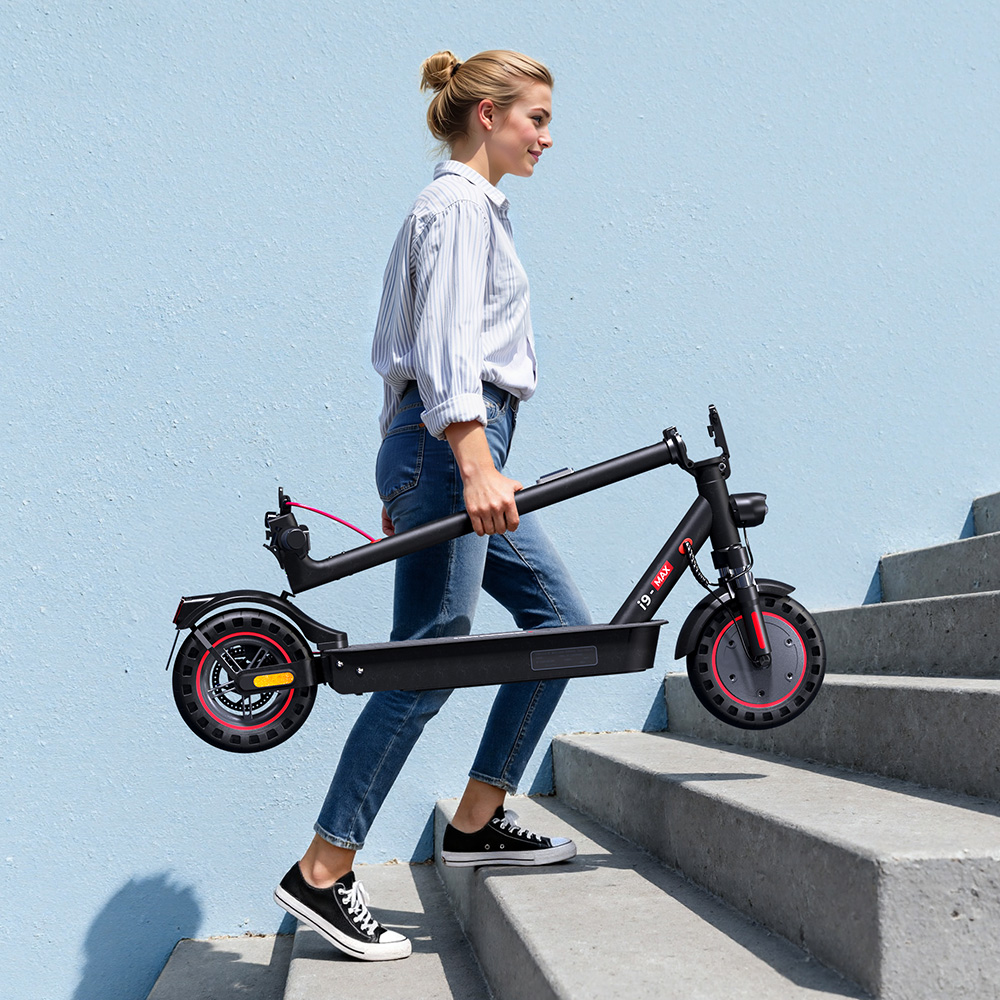 Una mujer lleva un monopatín eléctrico doblado subiendo escaleras frente a un muro azul.