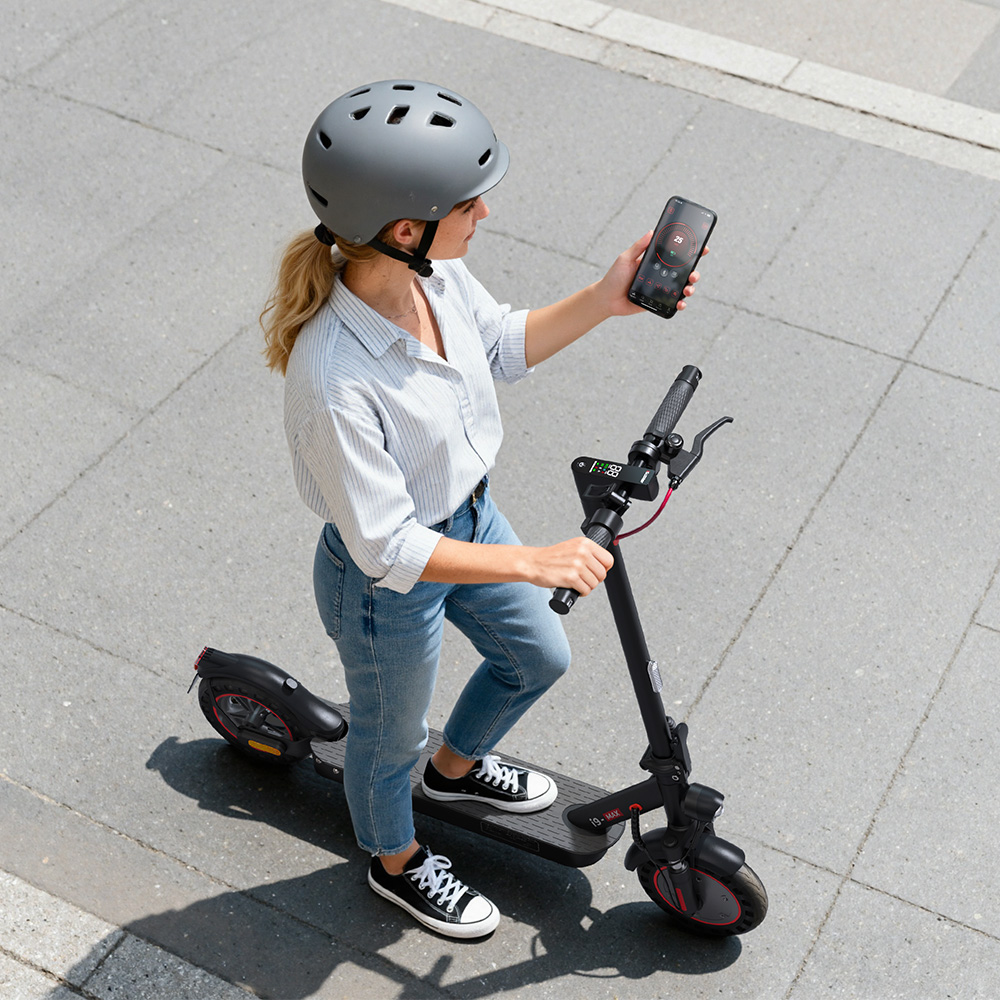 Mujer con casco y smartphone de pie sobre un patinete eléctrico negro en un suelo gris.