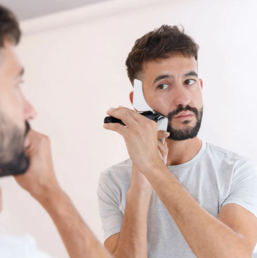 Un uomo con una maglietta grigia si rade la barba con un rifinitore elettrico davanti a uno specchio.