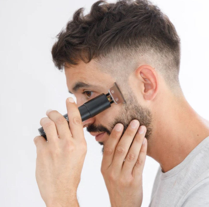 Un uomo con capelli scuri corti rifinisce la barba con un piccolo rifinitore nero e argento su sfondo bianco.